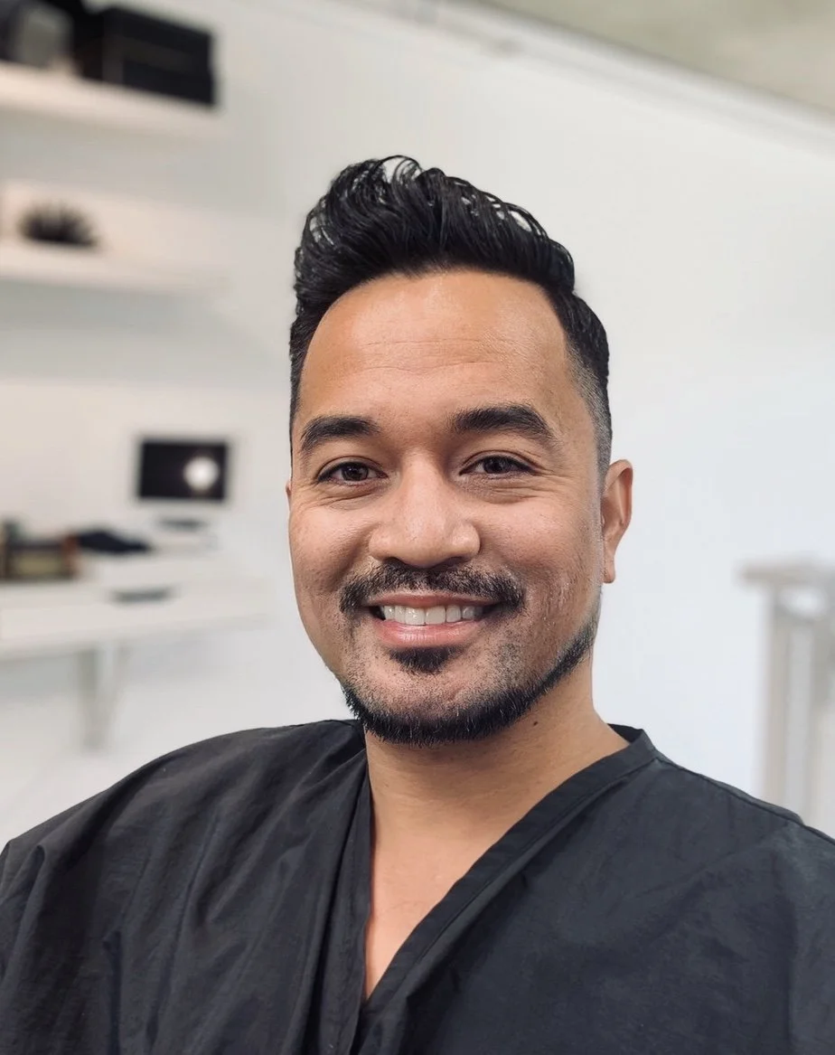 A smiling man taking a selfie in a room with white walls and shelves, dressed in black clothing.