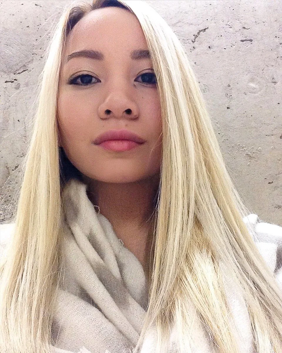 Close-up photo of a young woman with long blonde hair, fair skin, and minimal makeup, standing against a plain wall.