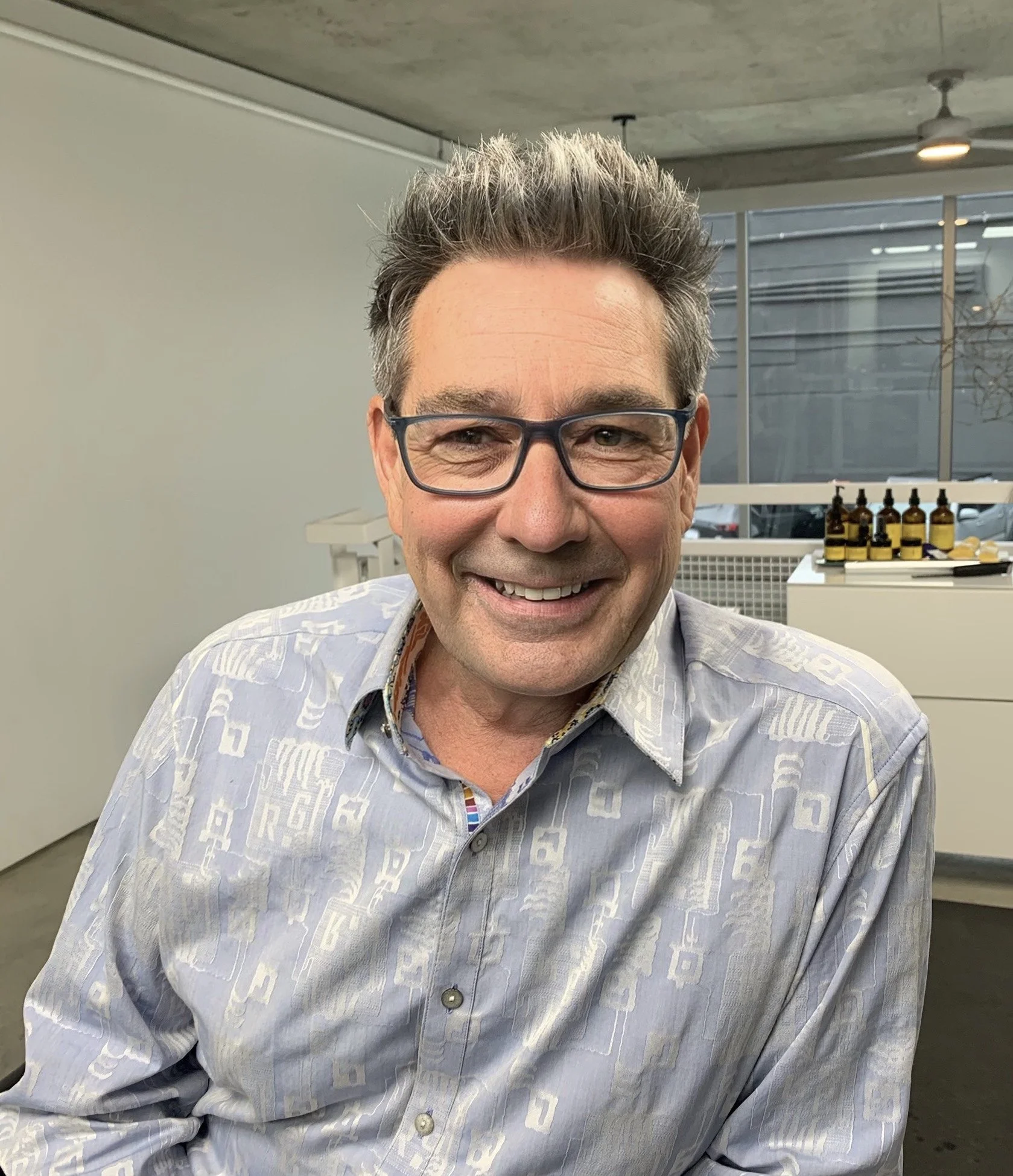 Smiling man with glasses and gray hair, wearing a patterned button-up shirt, in an indoor setting with bottles on a counter in the background.