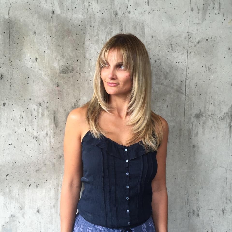 A woman with long blonde hair and bangs is standing against a gray concrete wall, wearing a navy blue sleeveless top and light blue patterned bottoms, looking to her left with a slight smile.
