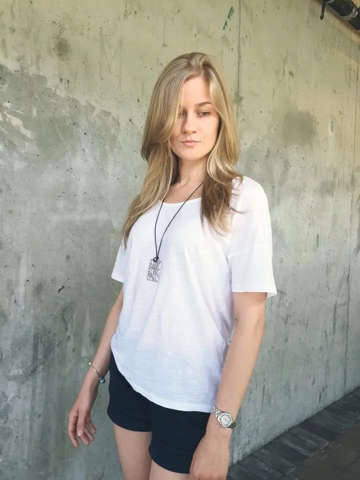 A young woman with long blonde hair wearing a white t-shirt, black shorts, and accessories, standing against a plain concrete wall.