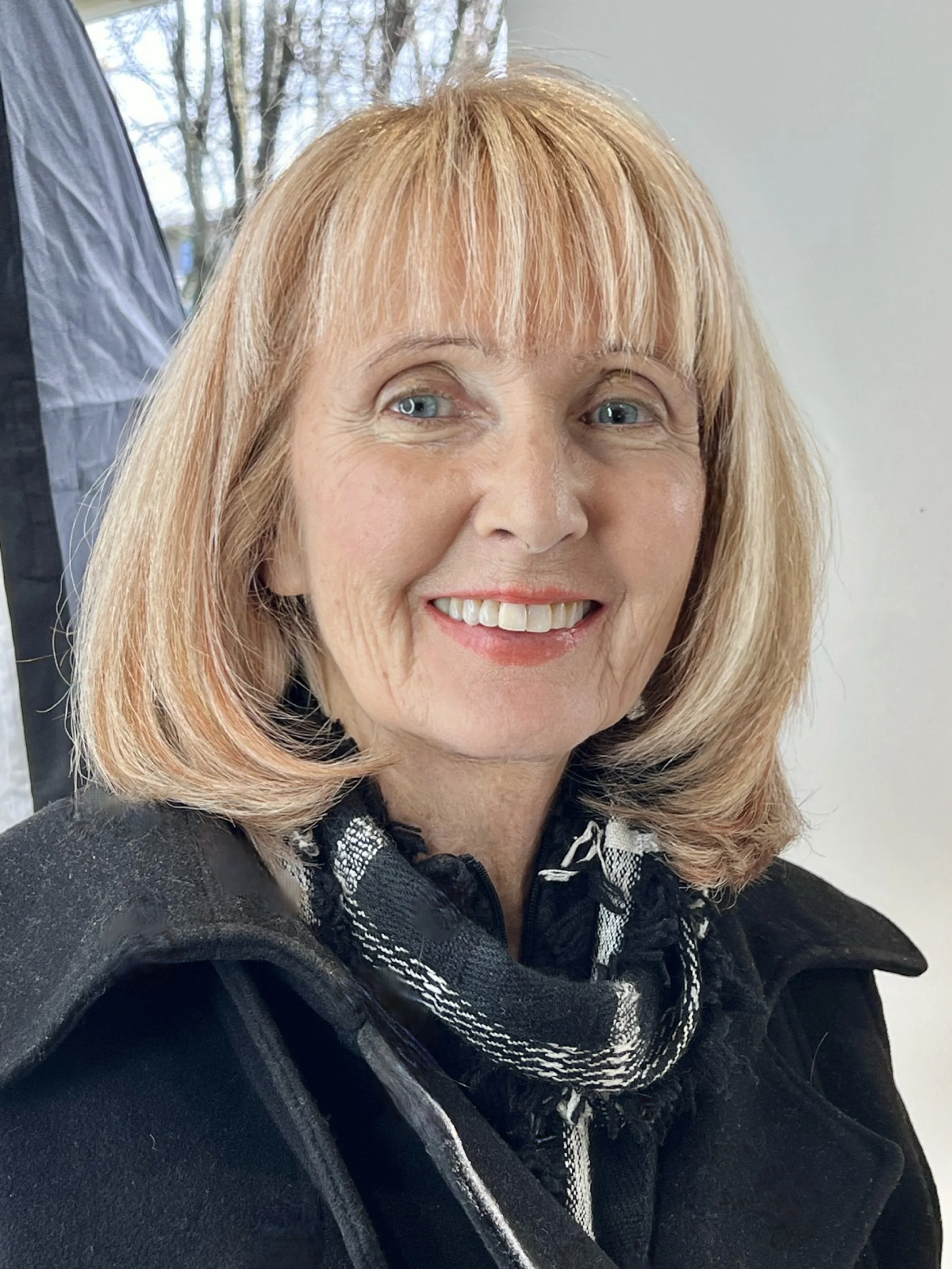Smiling woman with shoulder-length blonde hair, wearing a black coat and checkered scarf, against a blurred background.