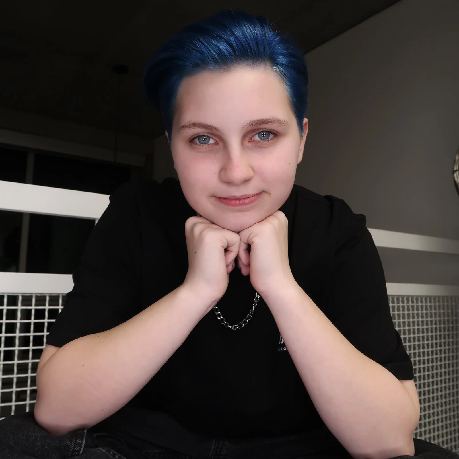 Person with blue hair and black shirt posing indoors, resting chin on hands.