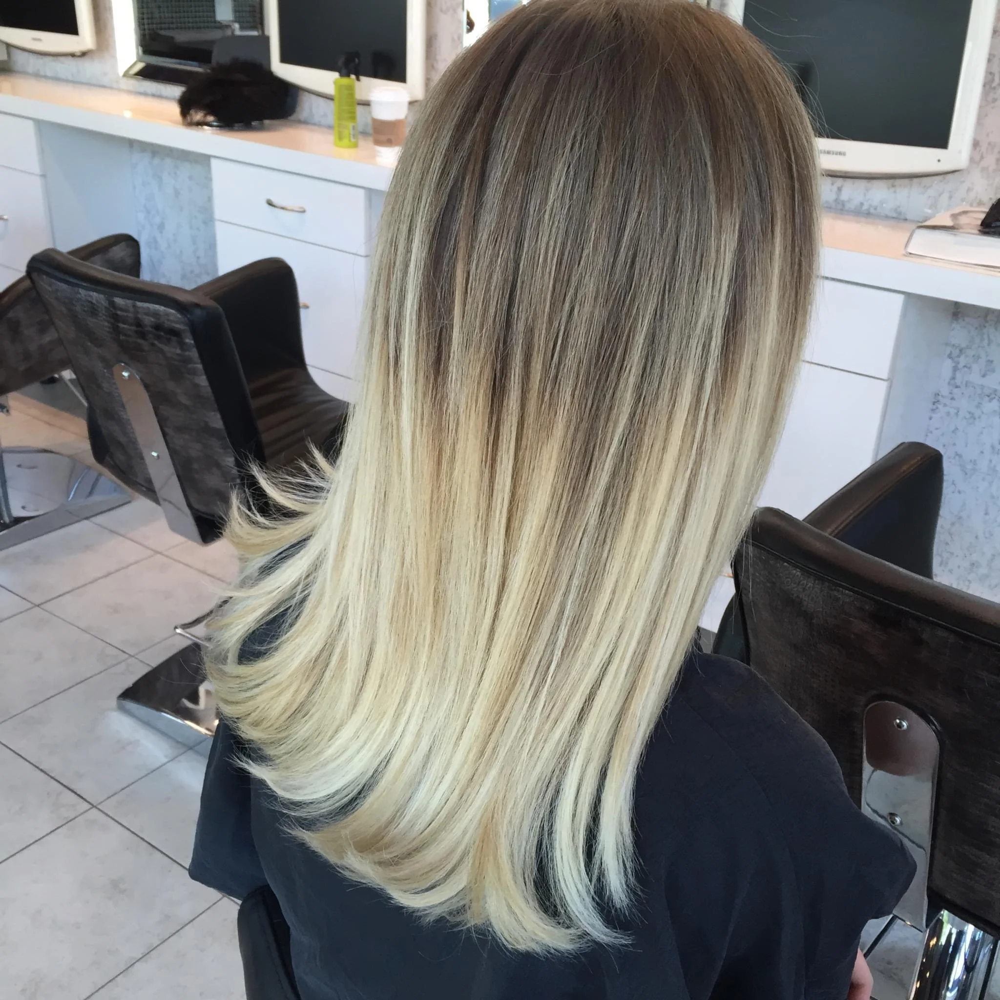 The image shows a woman with long, straight blonde hair with darker roots, sitting in a hair salon. The salon has black chairs, white counters, and several electronic devices, including monitors and a TV.