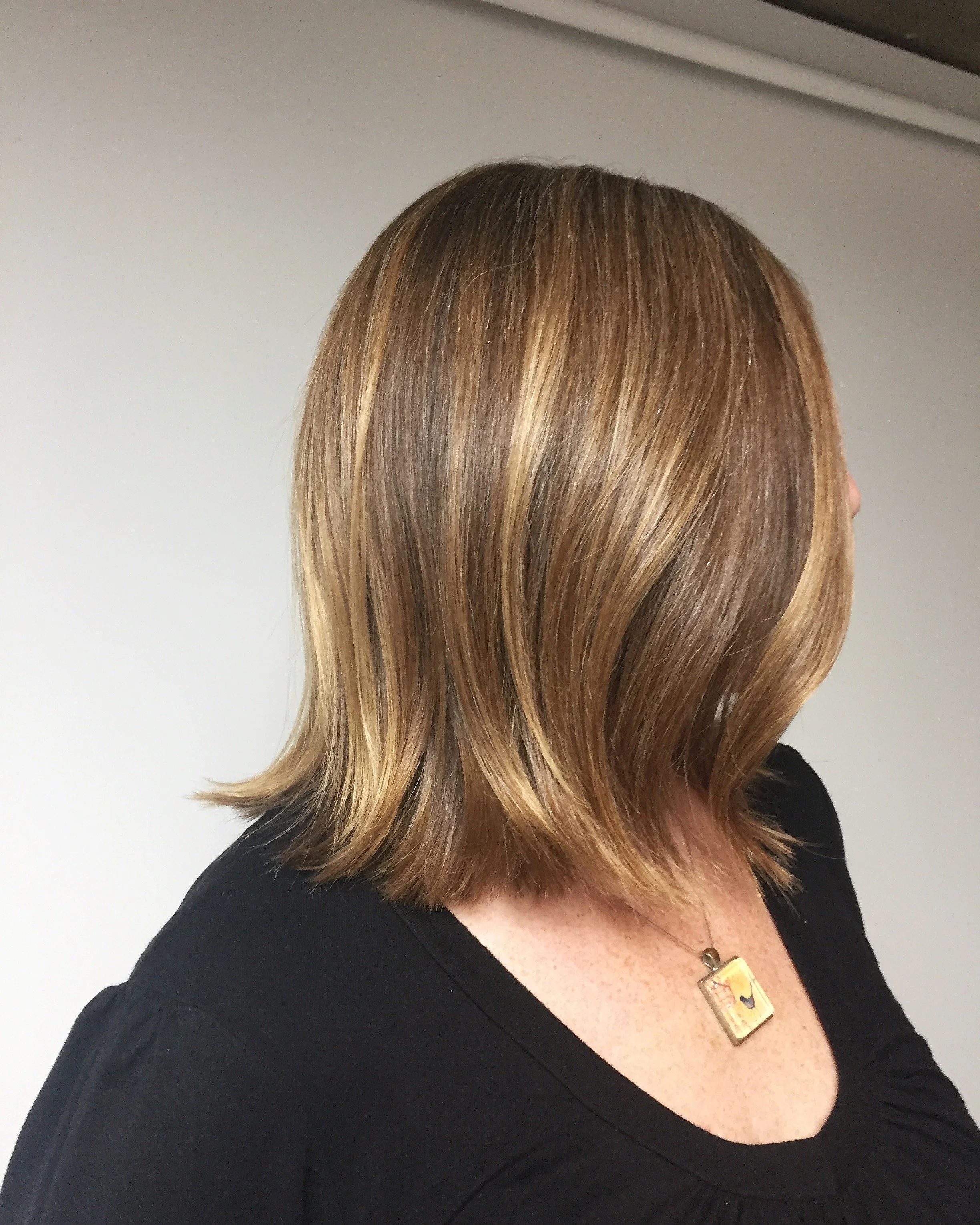 A woman with shoulder-length brown hair with highlights, wearing a black top and a pendant necklace, facing to the right against a plain background.