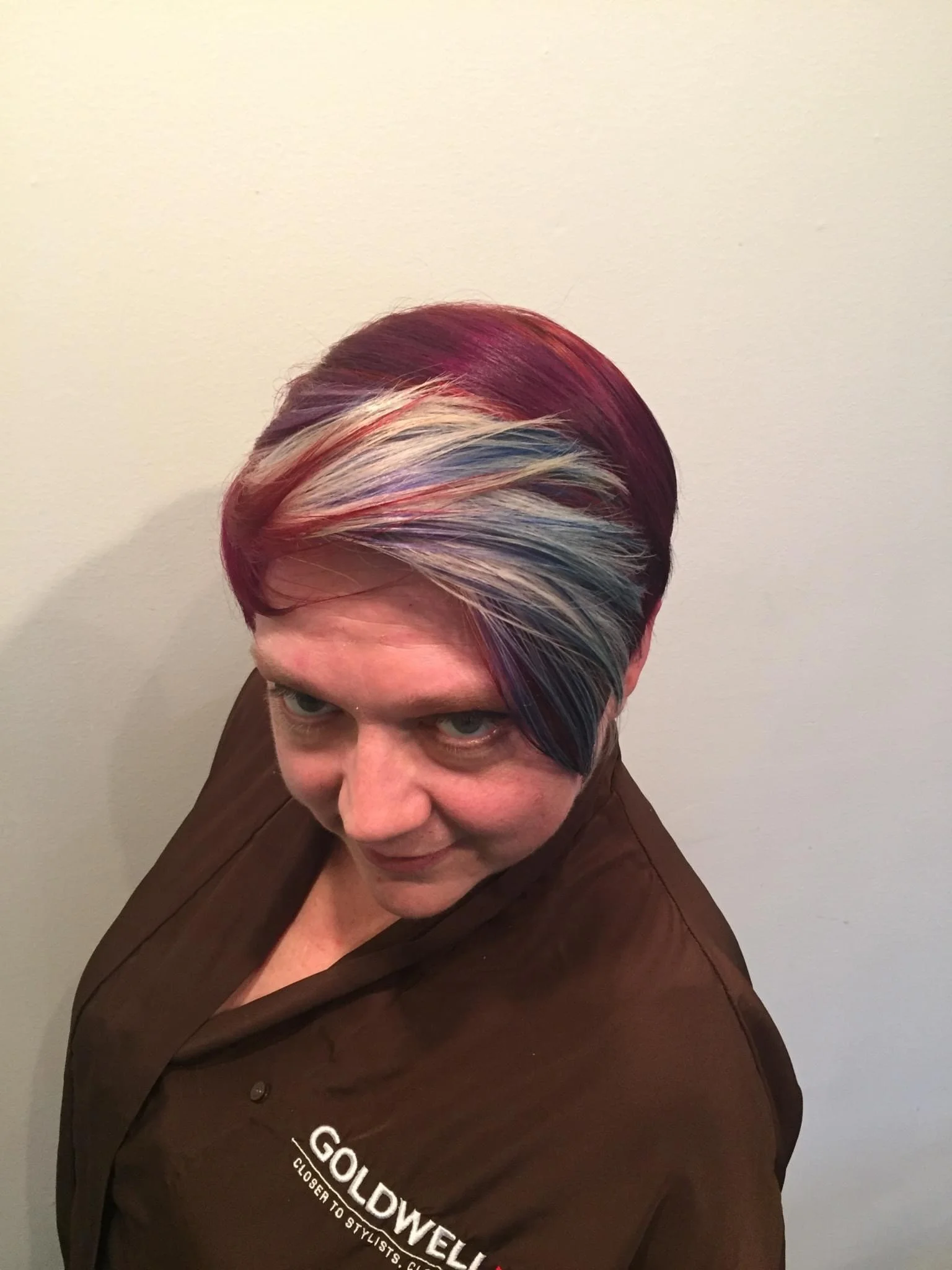 A woman with a colorful, layered hairstyle featuring shades of purple, pink, blue, and blonde, wearing a brown shirt with the GOLDFOWELL logo.