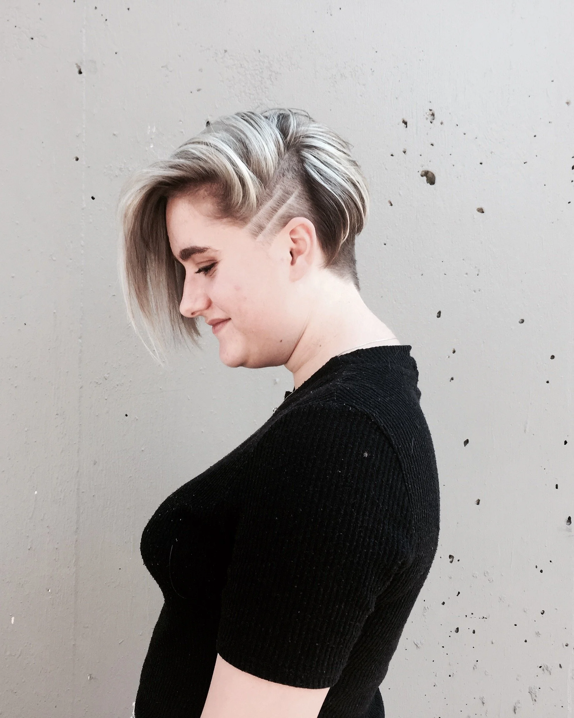 Side profile of a woman with an asymmetrical short hairstyle featuring shaved sides and longer top hair with blonde highlights, wearing a black top, standing against a plain wall.