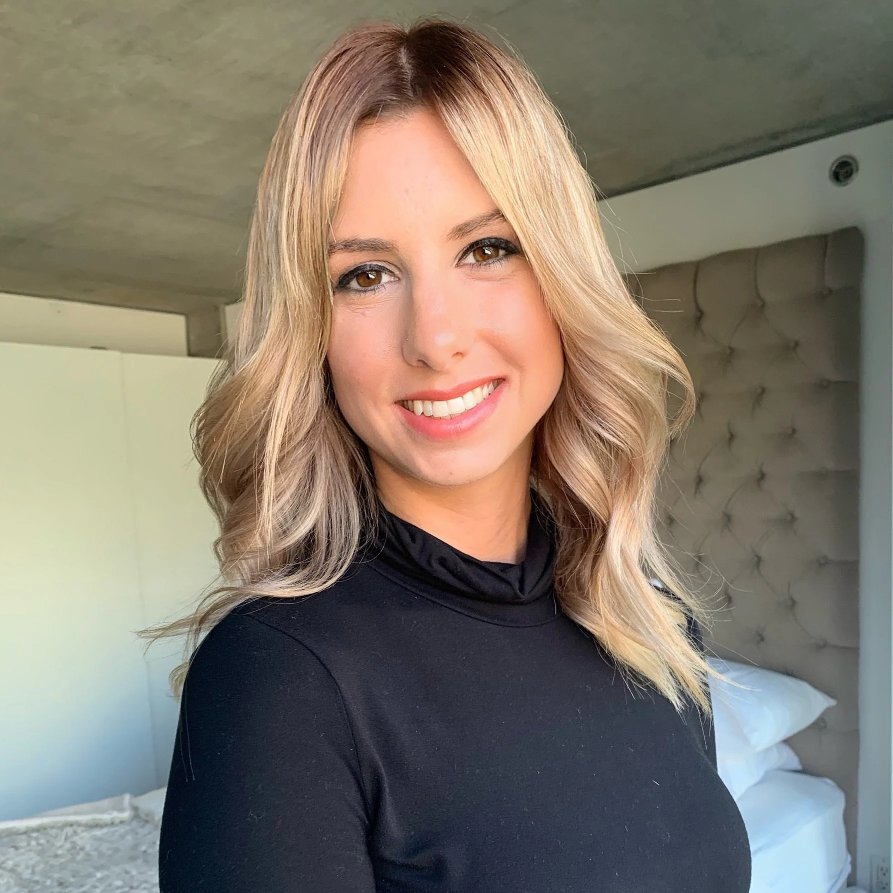 A smiling woman with blonde, wavy hair, wearing a black turtleneck in a bedroom with a tufted headboard and bed.