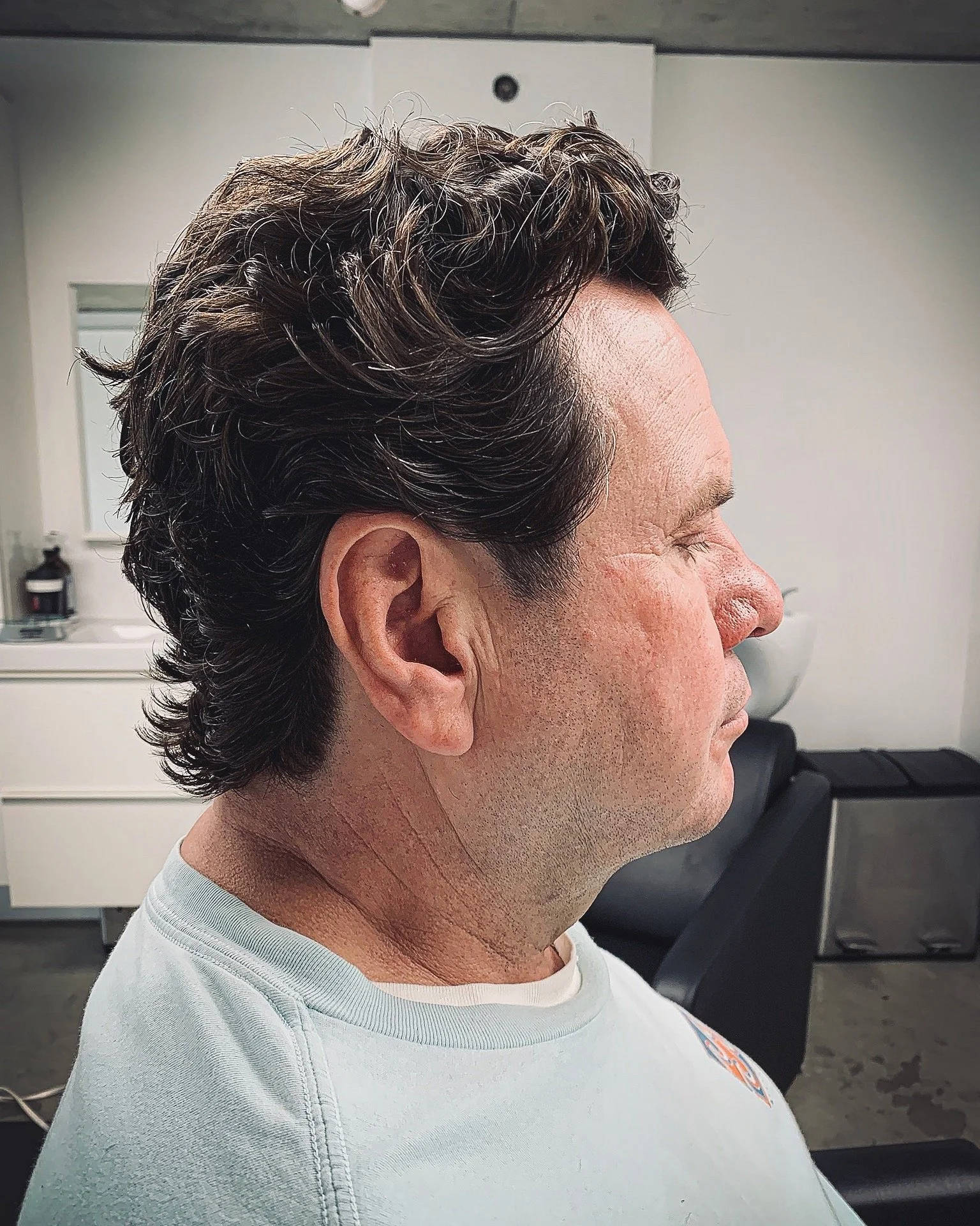 Side profile of a mature man with styled wavy dark hair sitting in a salon or barber shop.