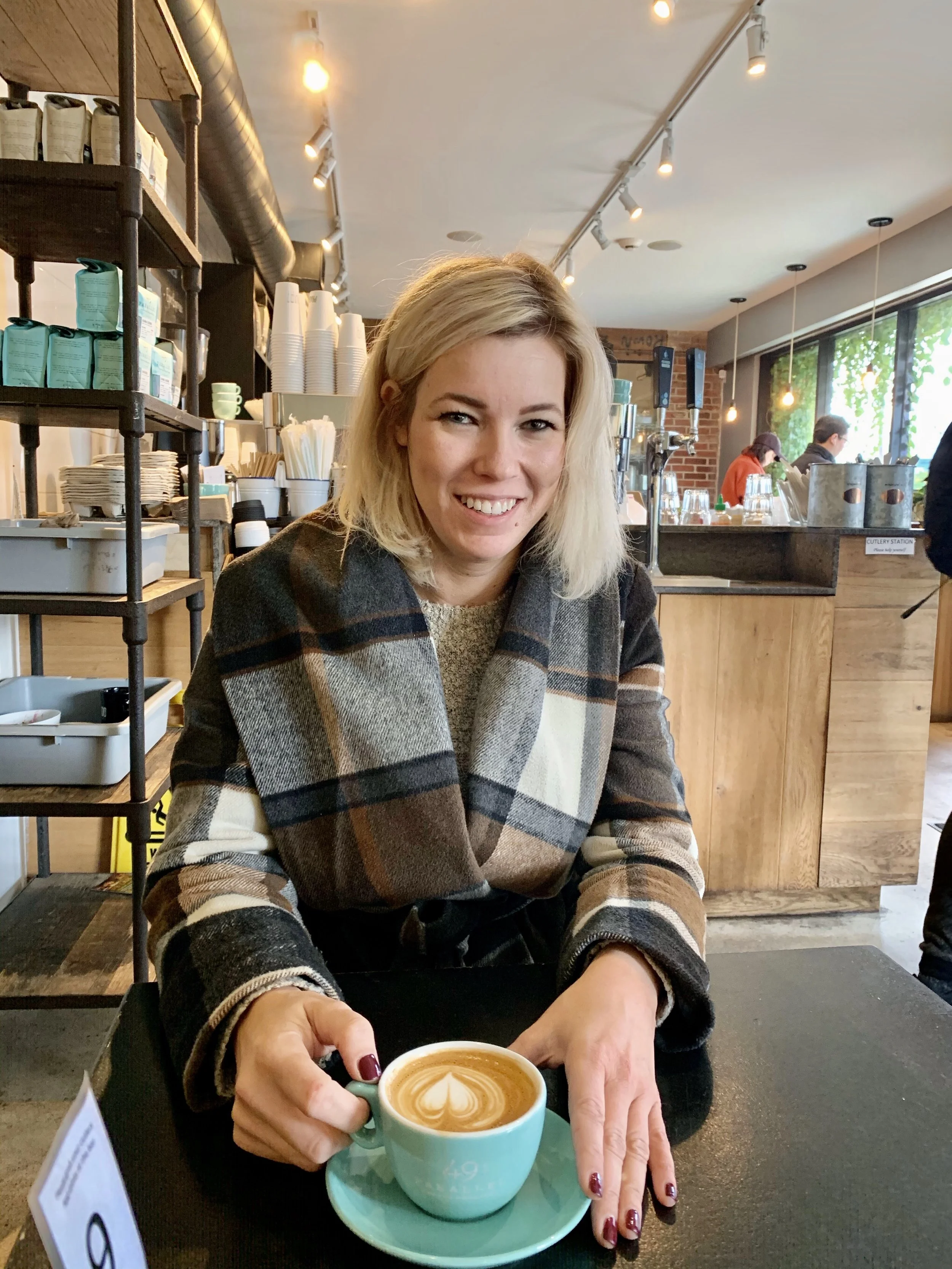 A woman with blonde hair smiling, sitting at a table in a coffee shop, holding a cup of coffee with latte art.
