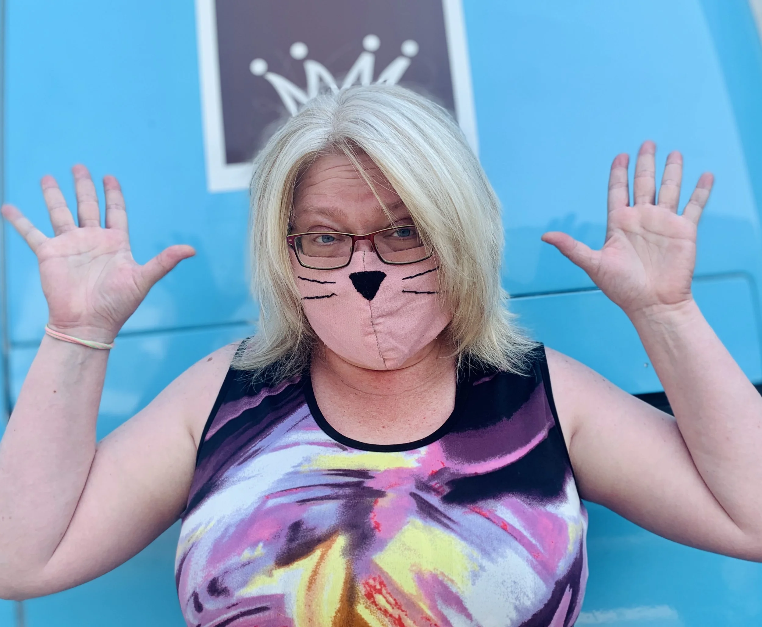 Person wearing a cat-themed face mask and colorful top, standing in front of a blue background with hands raised.