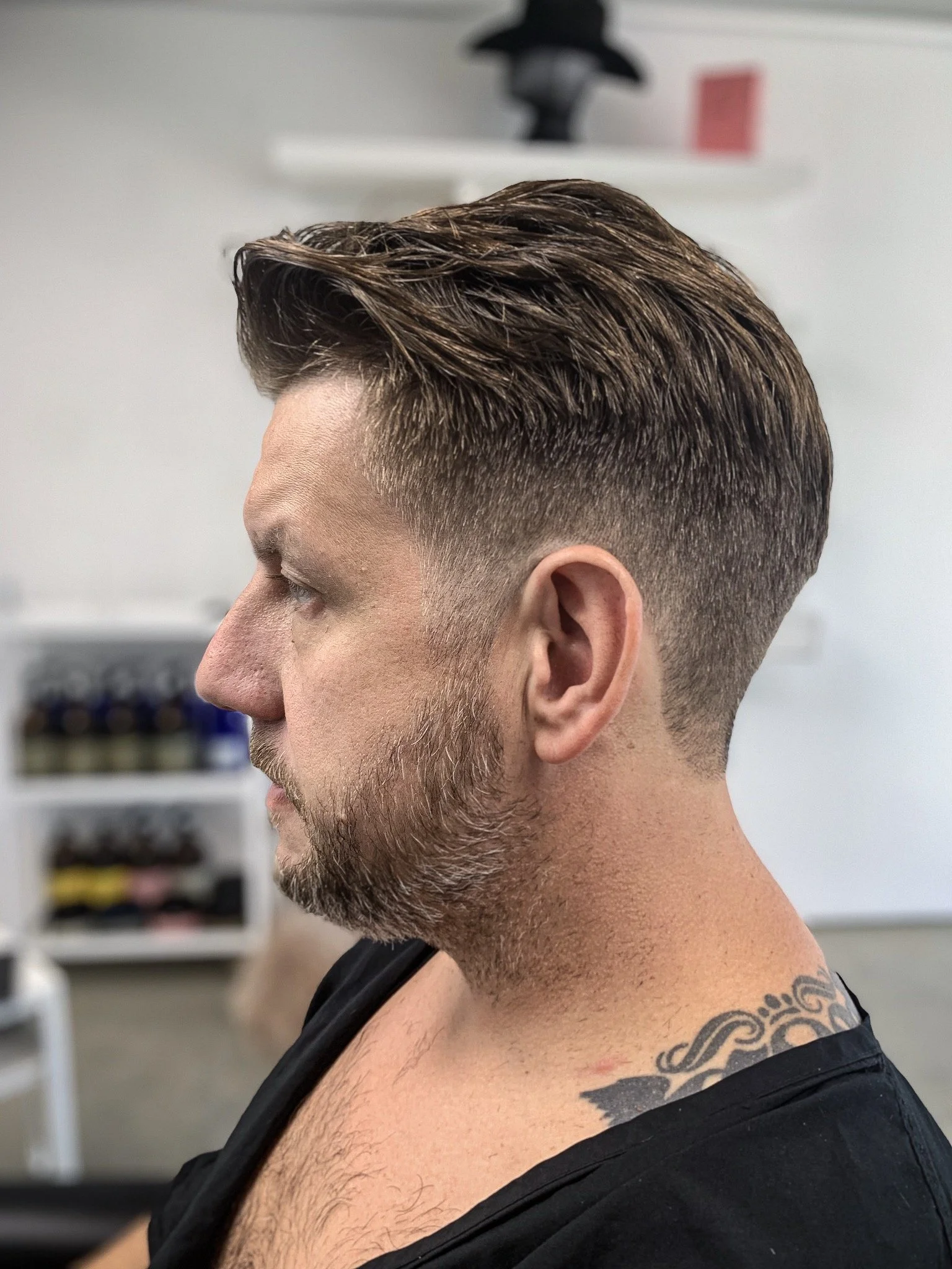 Side view of a man with a styled haircut and beard, featuring a neck tattoo, in an indoor setting.