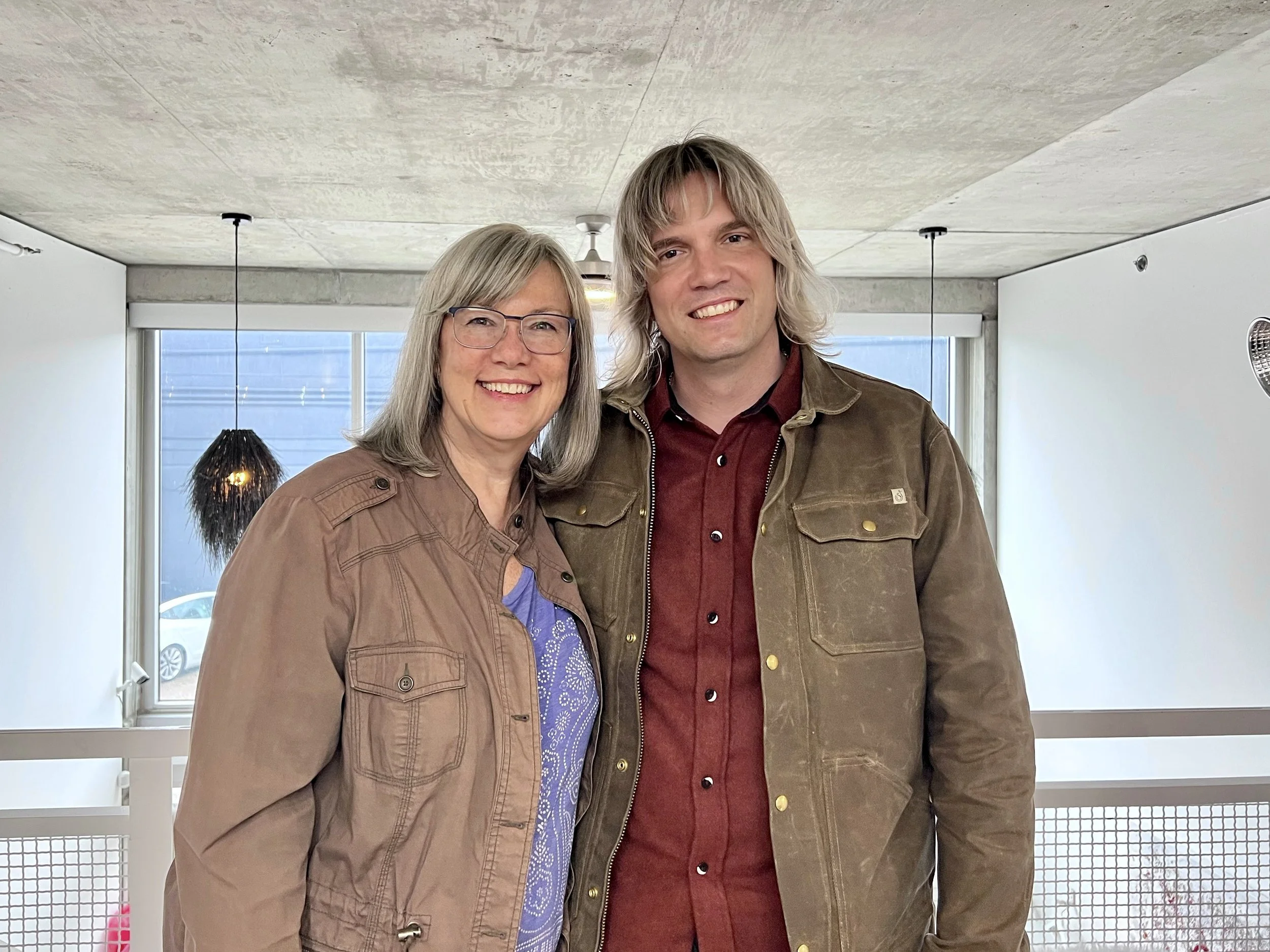 Two people smiling, standing indoors with modern decor, wearing casual jackets.