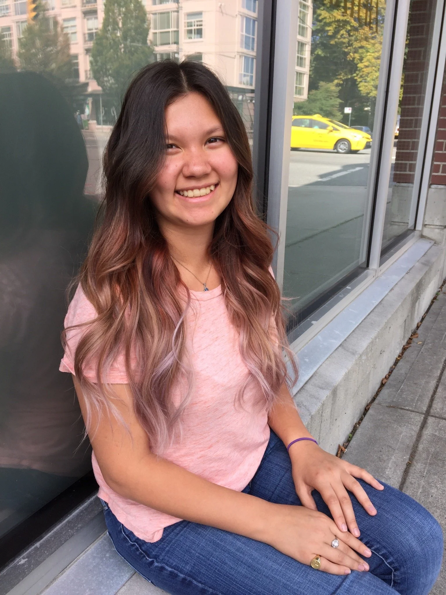 Young woman with long wavy hair sitting on a sidewalk, wearing a pink shirt and blue jeans, smiling. Background includes a building with windows and a yellow taxi passing by.