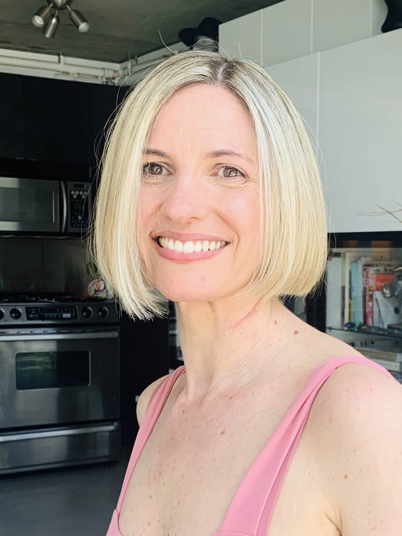 Smiling woman with short blonde hair in a kitchen, wearing a pink tank top.
