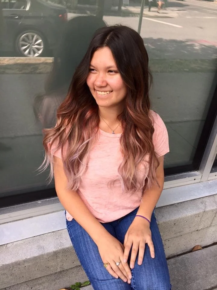 Smiling woman with long wavy hair sitting on a concrete ledge wearing a pink shirt and blue jeans, outdoors in front of a window reflection.