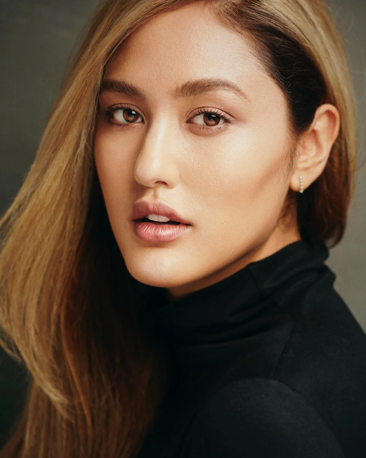 Close-up of a woman with long, straight hair, wearing a black turtleneck, with makeup, earrings, and a neutral background.