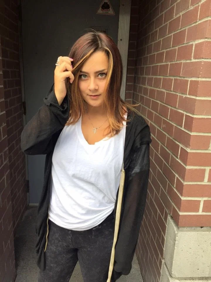 Young woman with brown and blonde hair standing between brick walls, touching her hair, wearing a white T-shirt, black jacket, and dark jeans.