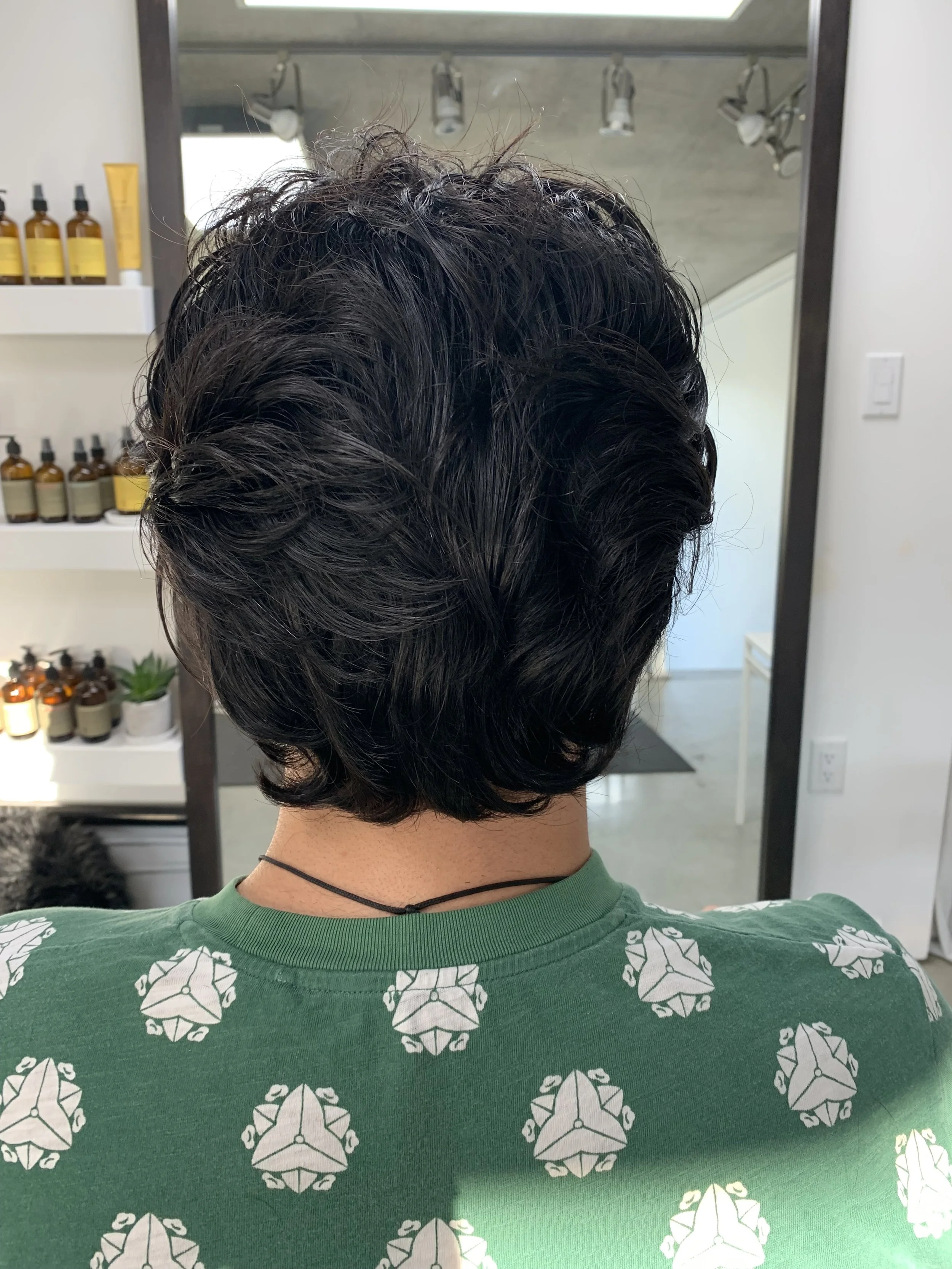 A person with short, wavy black hair sitting in front of a mirror in a salon, wearing a green shirt with a white geometric pattern and a black choker necklace. Hair products are visible on shelves to the left.