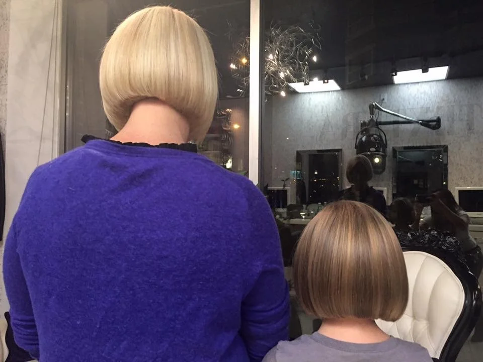 Back view of a woman with a short blonde bob haircut wearing a blue sweater, sitting next to a young girl with a similarly styled short blonde bob haircut, in a room with people and a recording studio in the background.