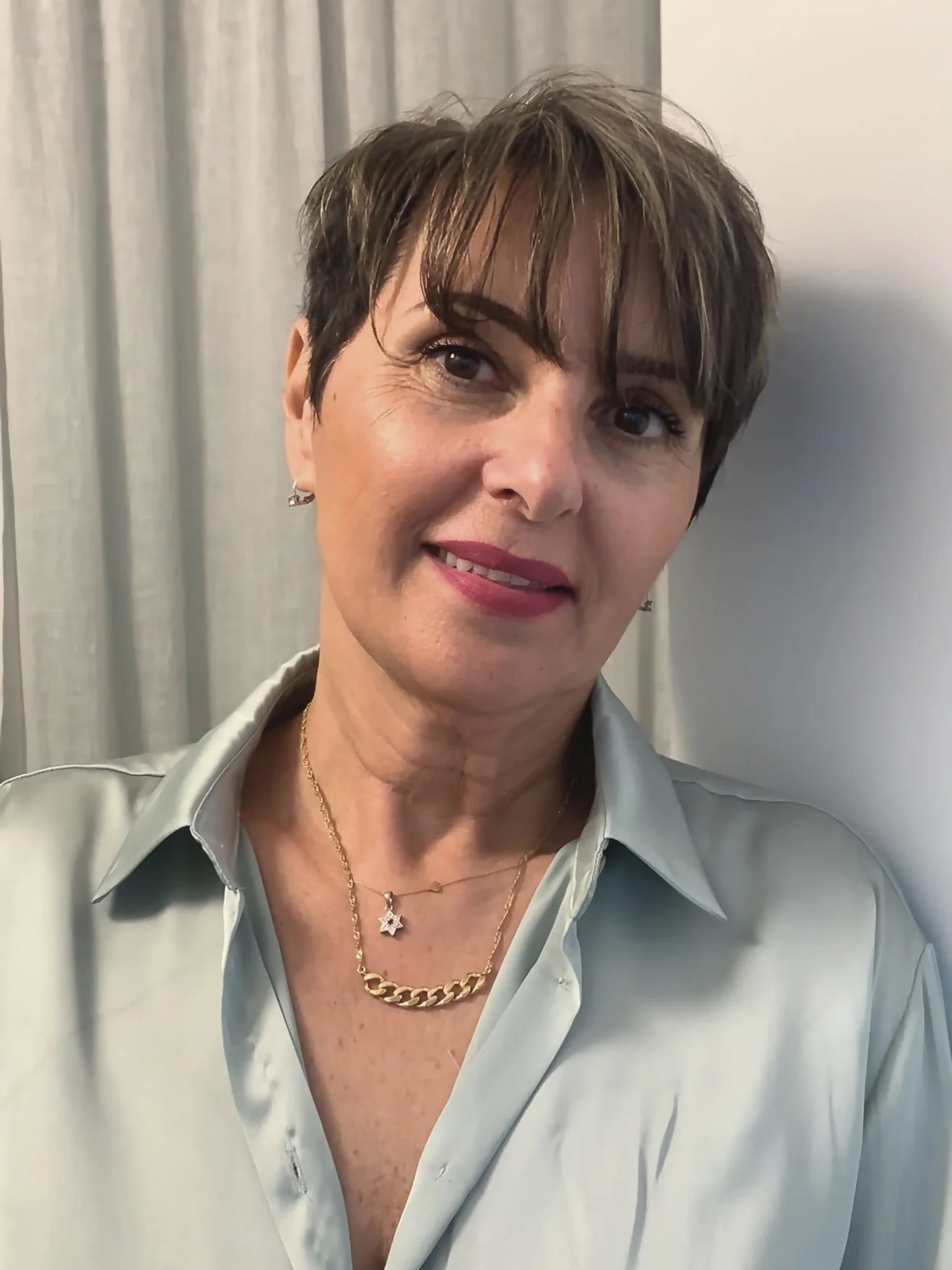 A woman with short brown hair wearing a light blue blouse and layered gold necklaces, standing against a light-colored background.