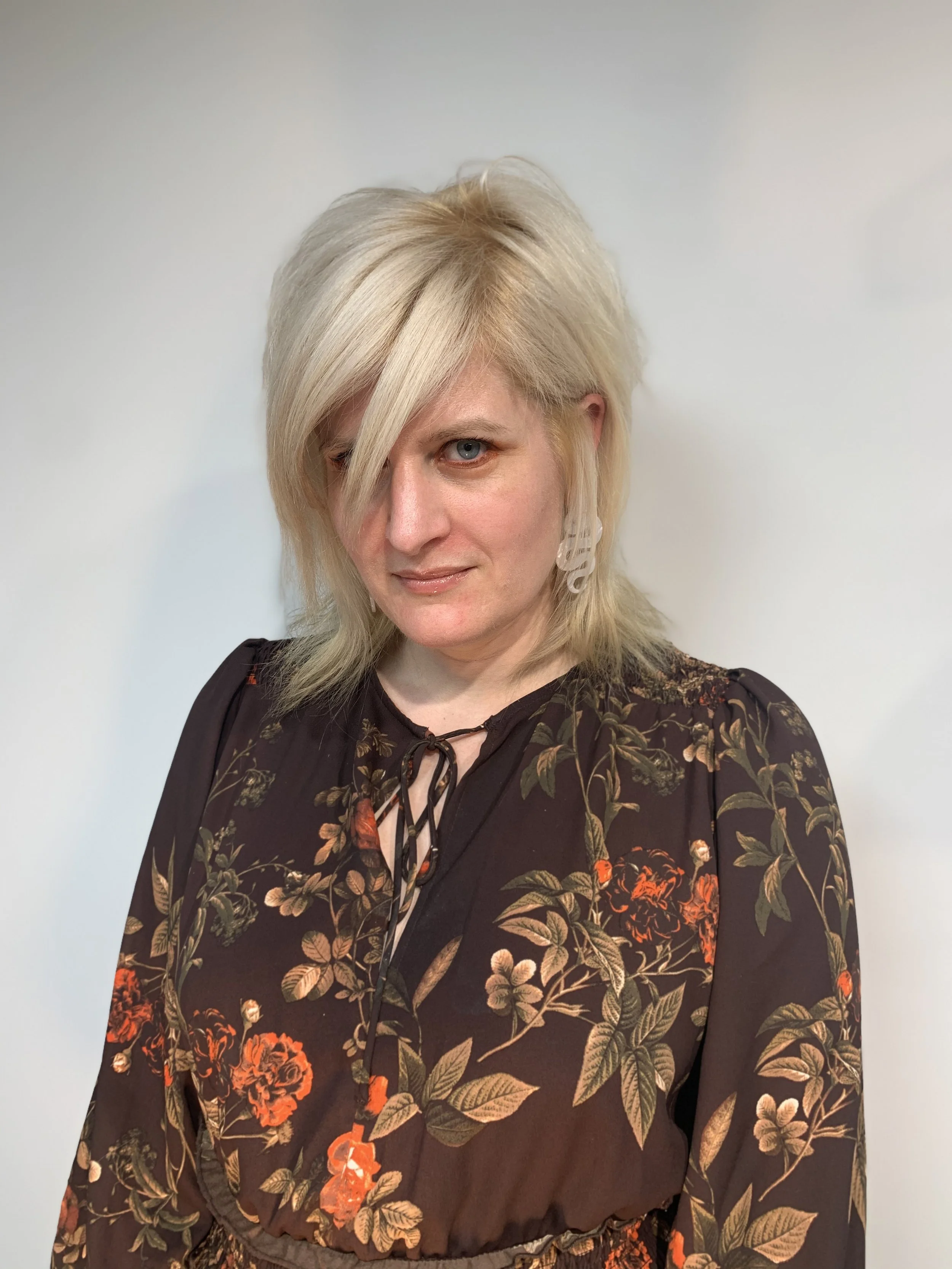 A woman with platinum blonde, layered hair, partly covering her face, wearing a black dress with a floral pattern in red, orange, beige, and green, standing against a plain white background.