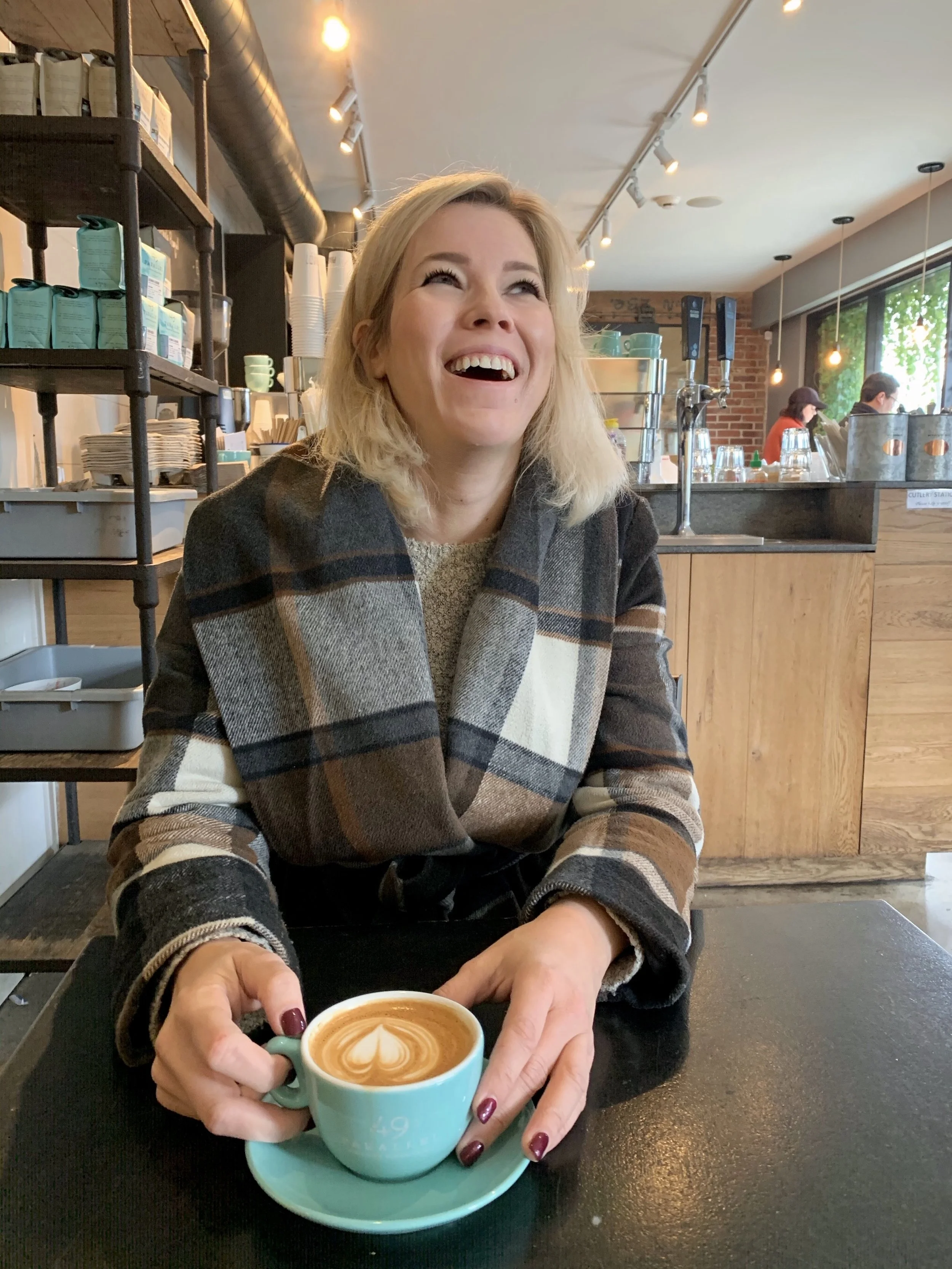 A woman sitting at a cafe table, holding a turquoise coffee cup with latte art, smiling and looking up.