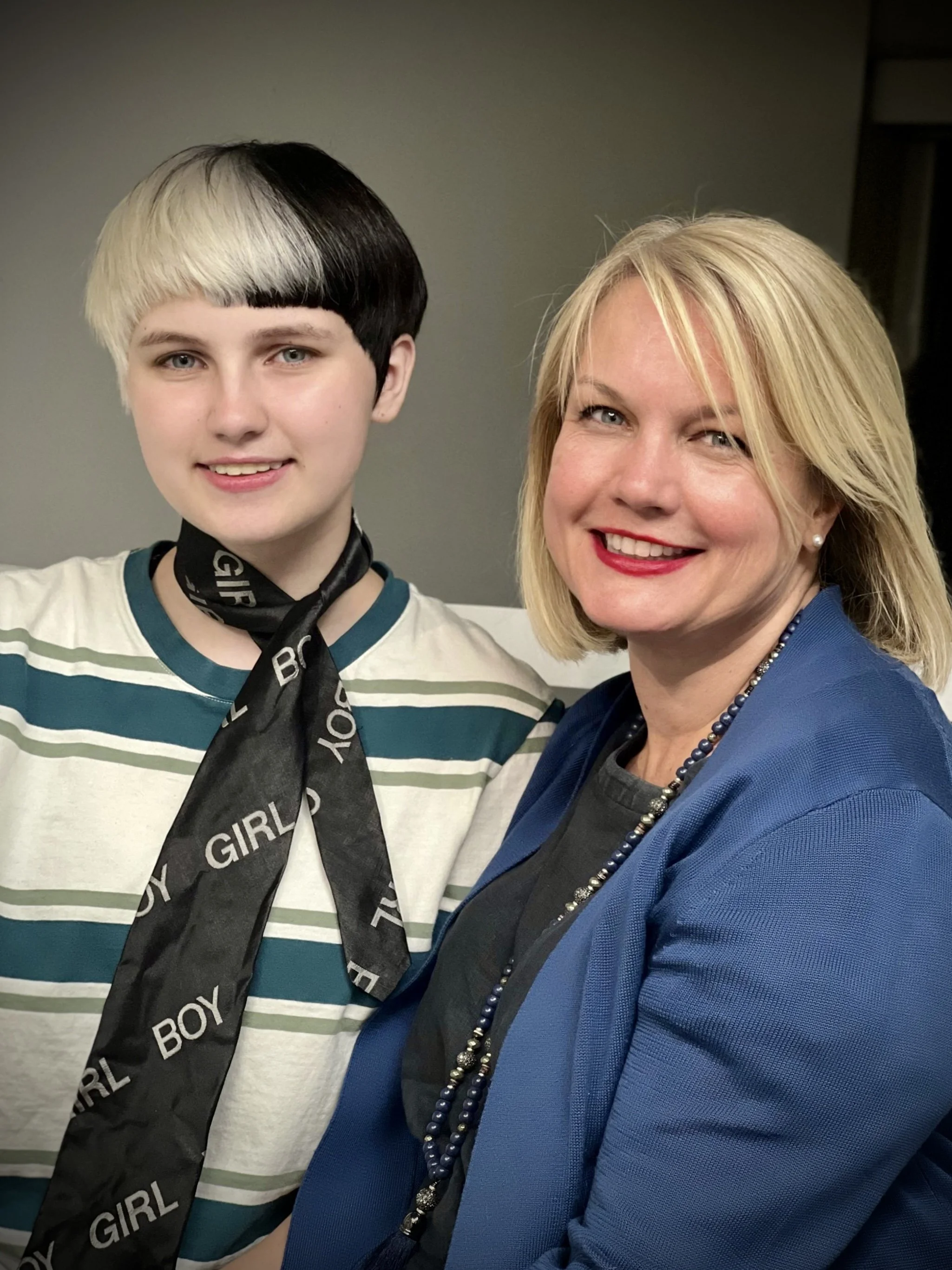 Two people smiling, one with a black and white bob haircut and a striped shirt and scarf, the other with blonde hair and a blue blazer.