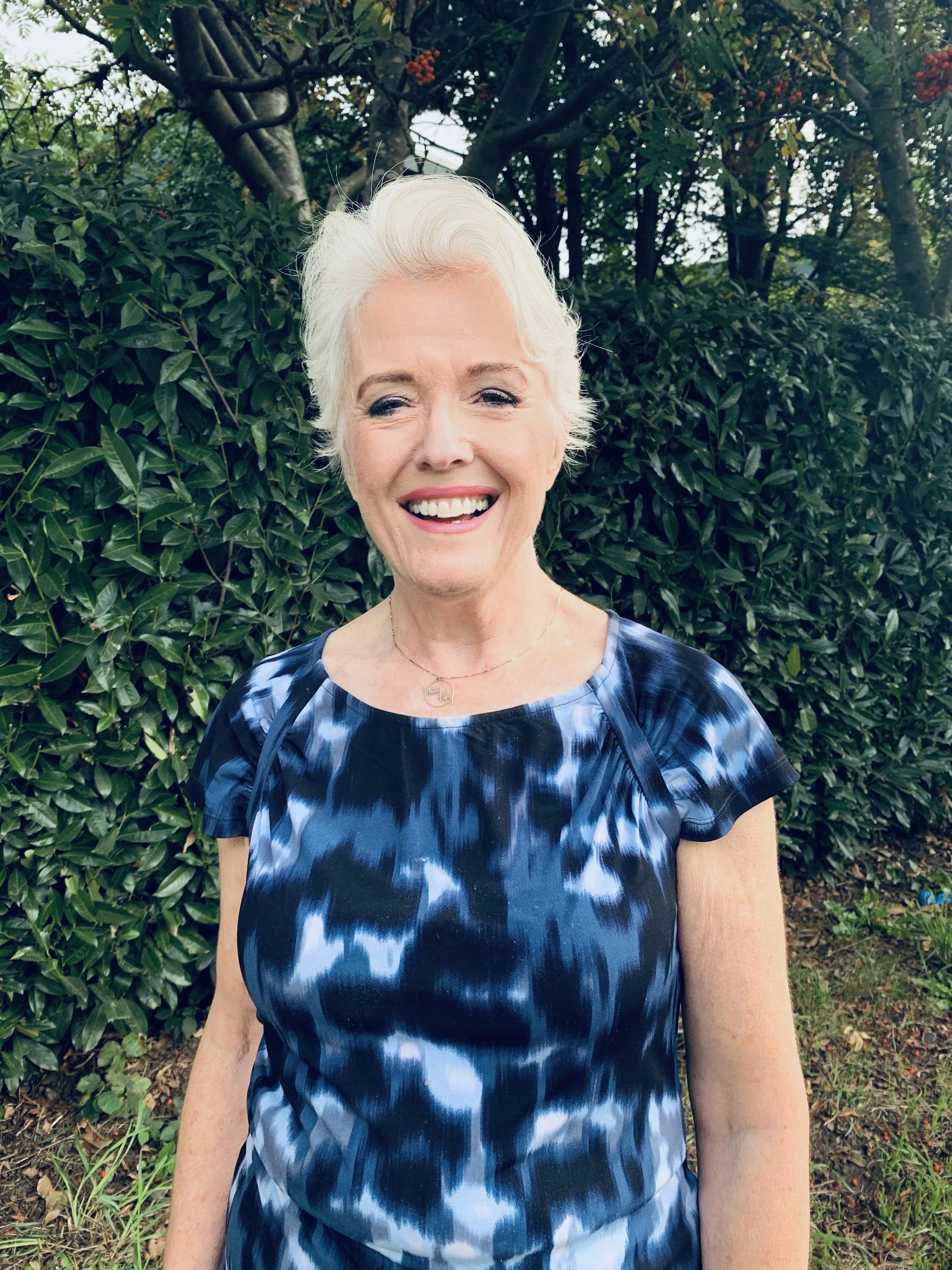 A smiling woman with short white hair standing outdoors in front of a leafy green hedge, wearing a blue and black tie-dye dress and a gold necklace.