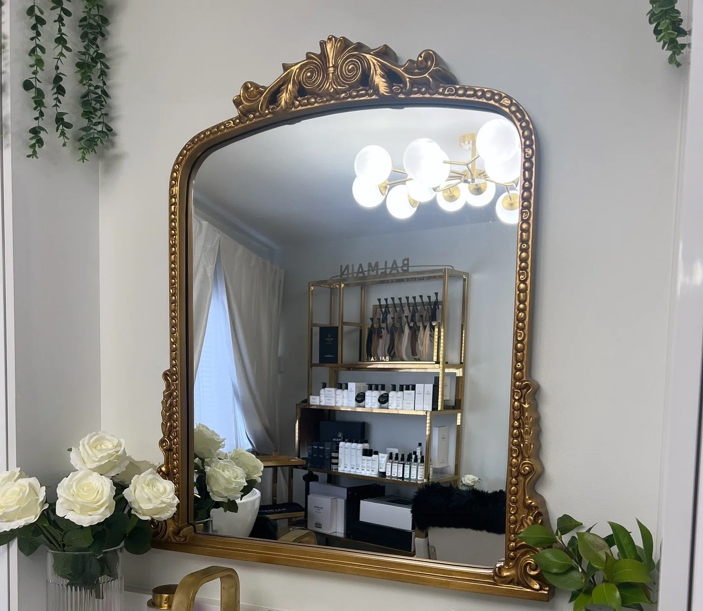 Decorative mirror with a gold ornate frame reflecting a modern chandelier, shelves stocked with skincare or beauty products, and a 'BALMAIN' sign in a stylish salon or boutique display.