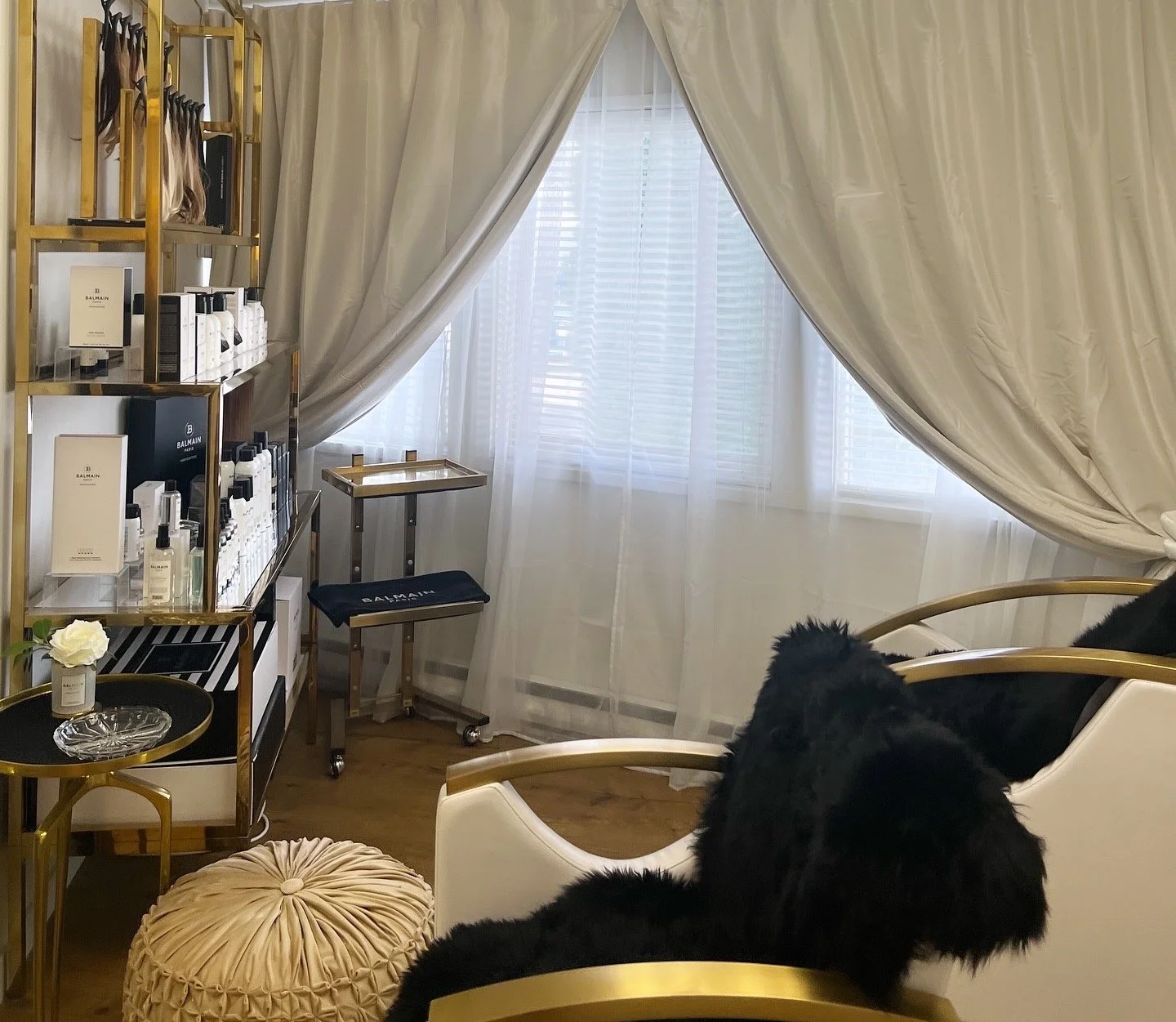 A luxurious room with a gold and white color scheme. It features a white sofa with black pillows, a gold side table with a flower and decorative glass dish, a gold shelving unit with skincare or cosmetic products, a small rolling cart, sheer curtains, and a window with blinds.