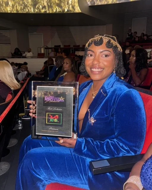 Woman sitting in theater holding award plaque, wearing a blue velvet suit with gold braid accents, smiling at camera.