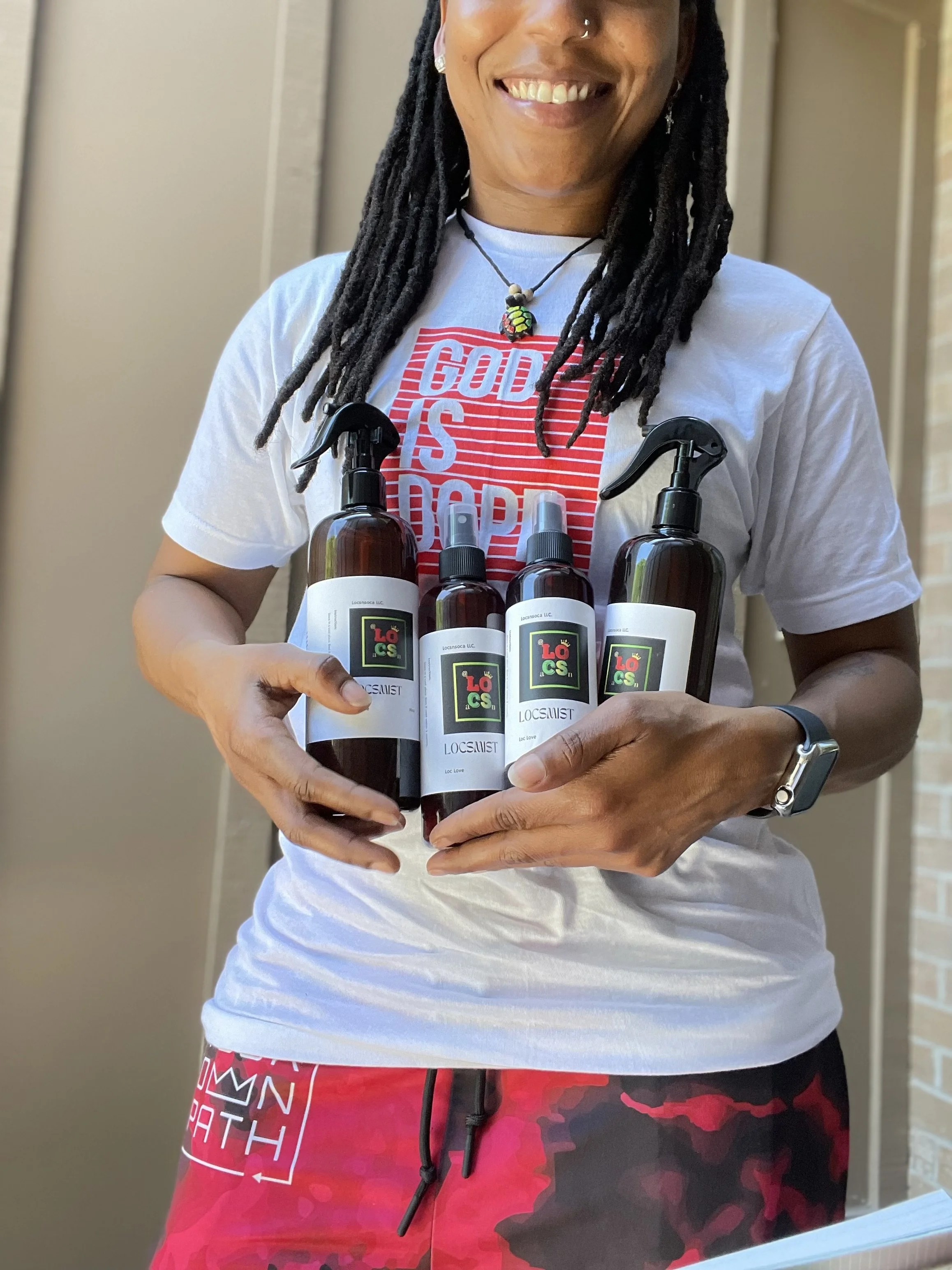 Person with dreadlocks holding four bottles of spray or lotion, standing outdoors near a beige wall, smiling.