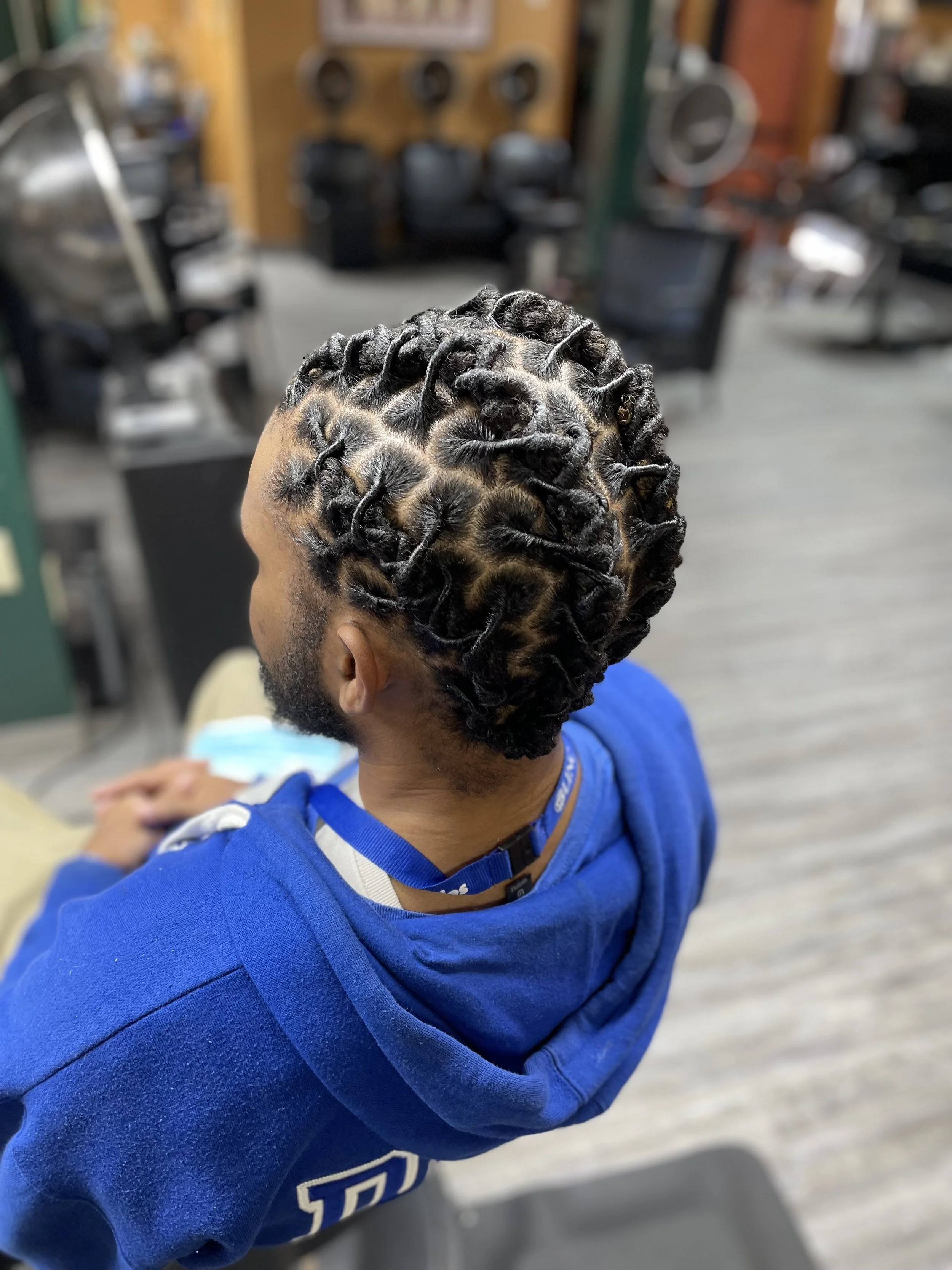 A man with black dreadlocks styled in a circular pattern, sitting in a barbershop, wearing a blue hoodie.