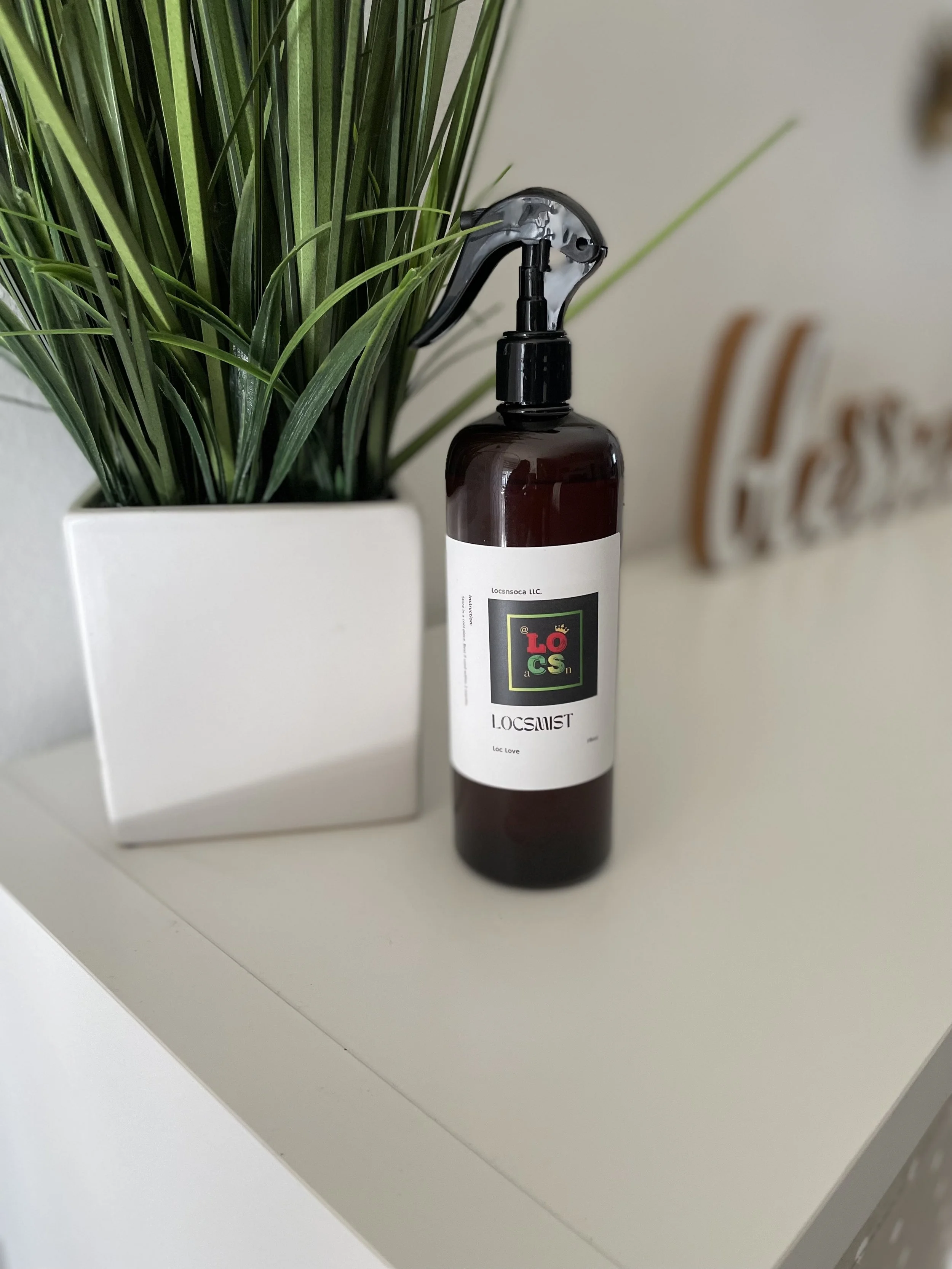 A spray bottle labeled 'LOCSMIST' leaning against a potted green plant on a white surface.