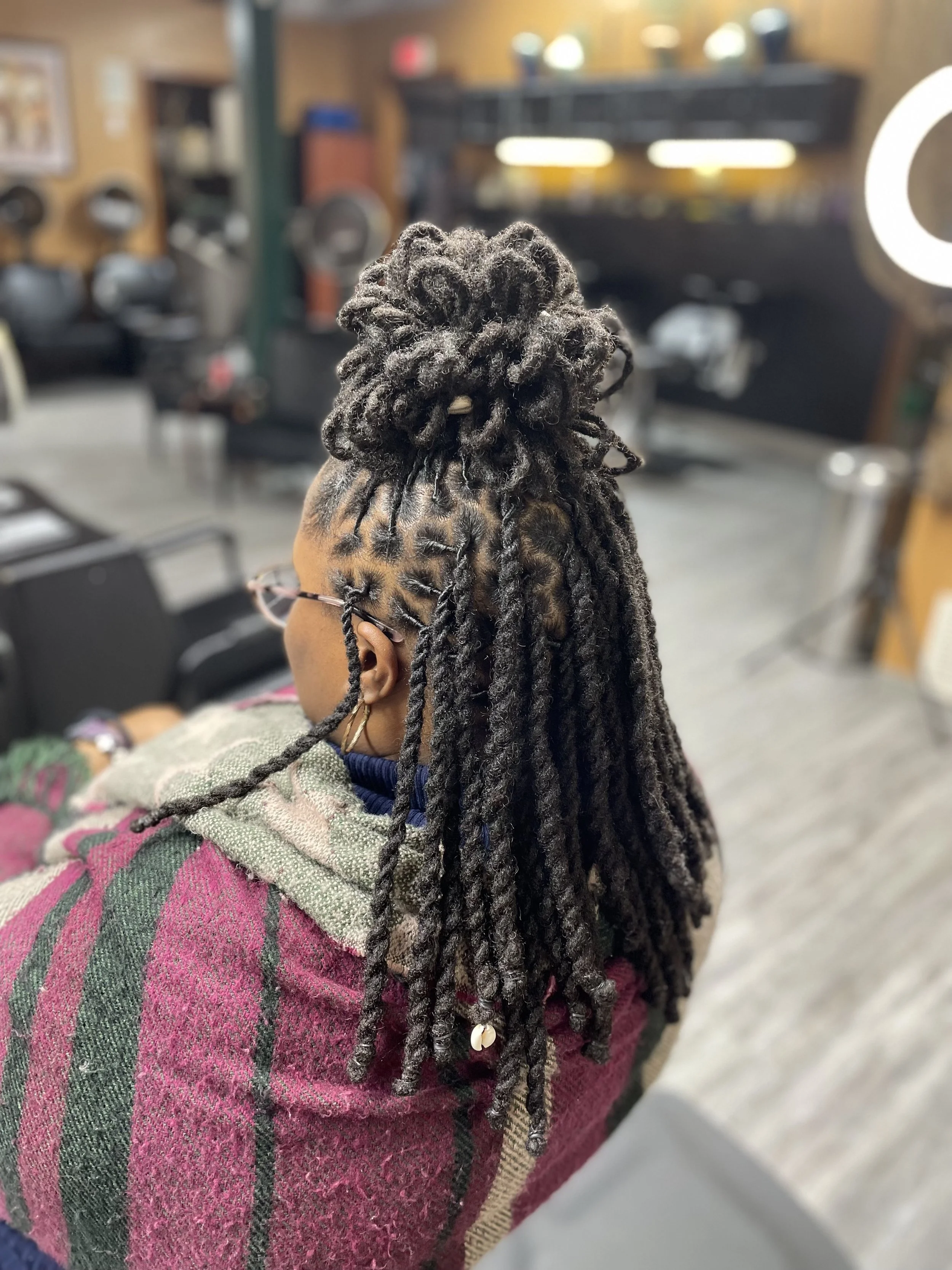 A woman with styled dreadlocks tied in a top bun, wearing glasses and hoop earrings, seated in a hair salon with blurred background.