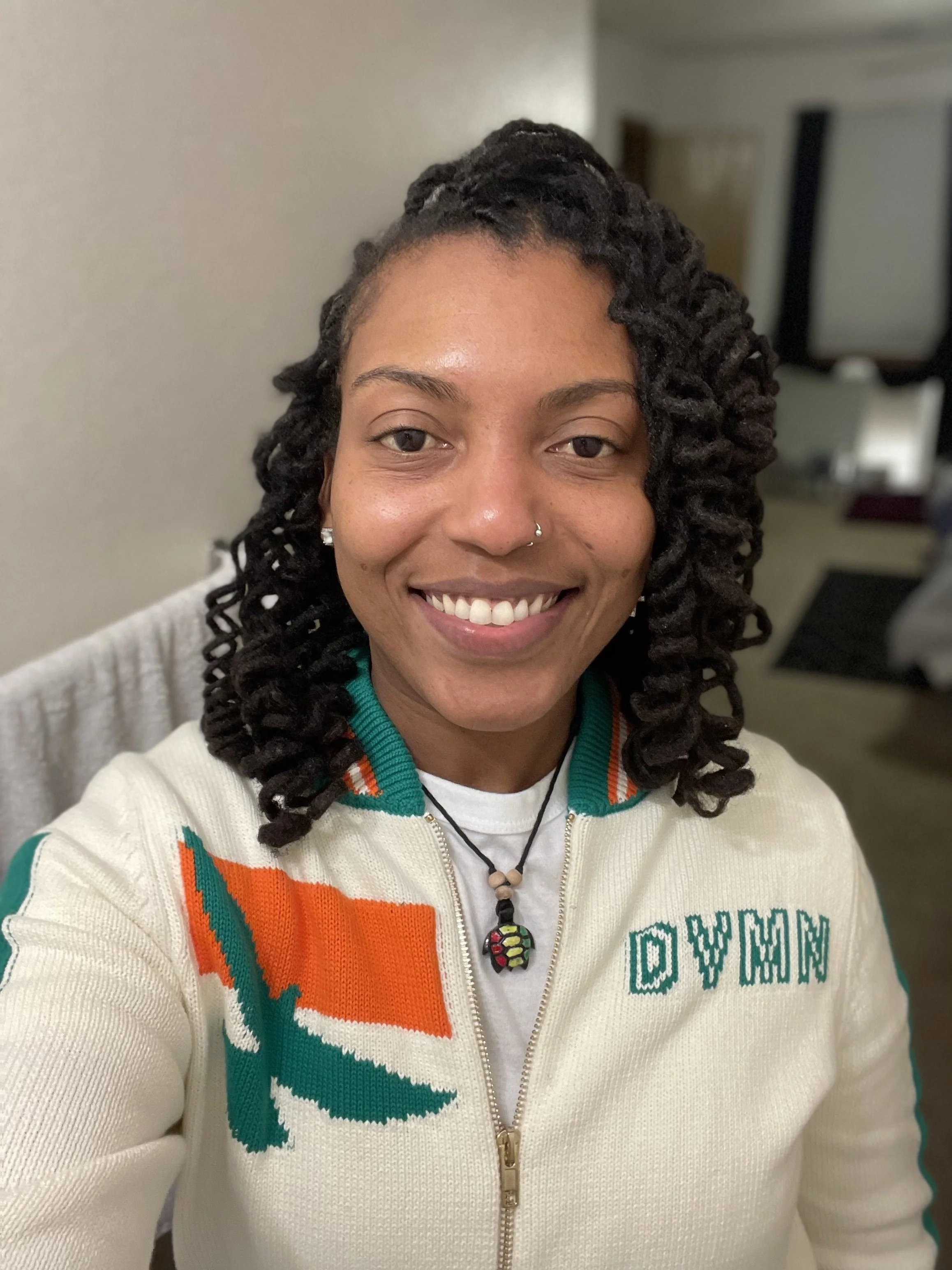 A woman smiling for a selfie, wearing a cream-colored sweater with orange and green accents, and the word 'WOMAN' embroidered on it. She has dark, styled hair, a nose piercing, and a colorful beaded necklace.