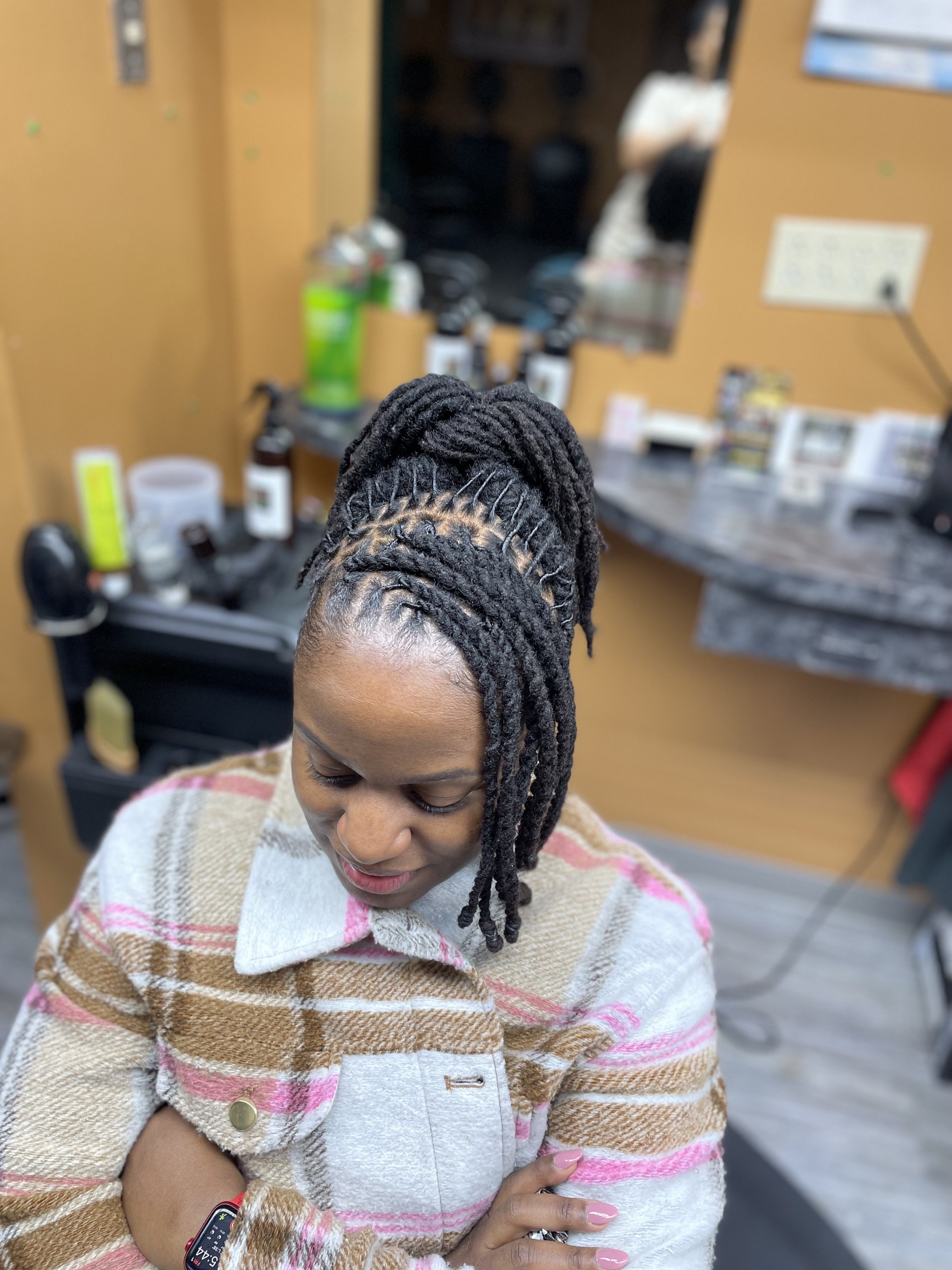 A woman with styled dreadlocks sitting in a hair salon chair, looking downward with her arms crossed, wearing a beige plaid jacket.