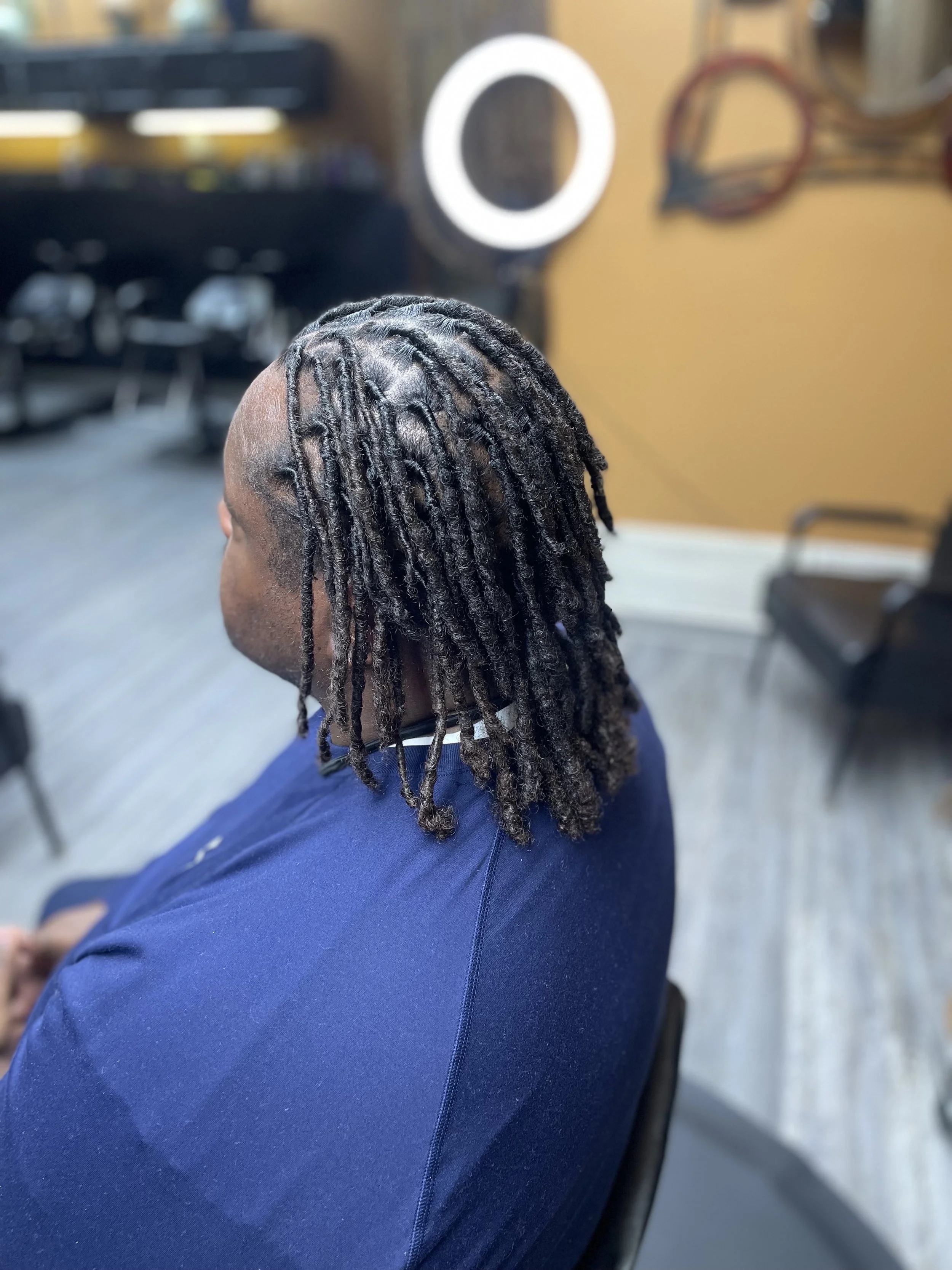 Side view of a man with short dreadlocks sitting in a salon chair, wearing a dark blue shirt.