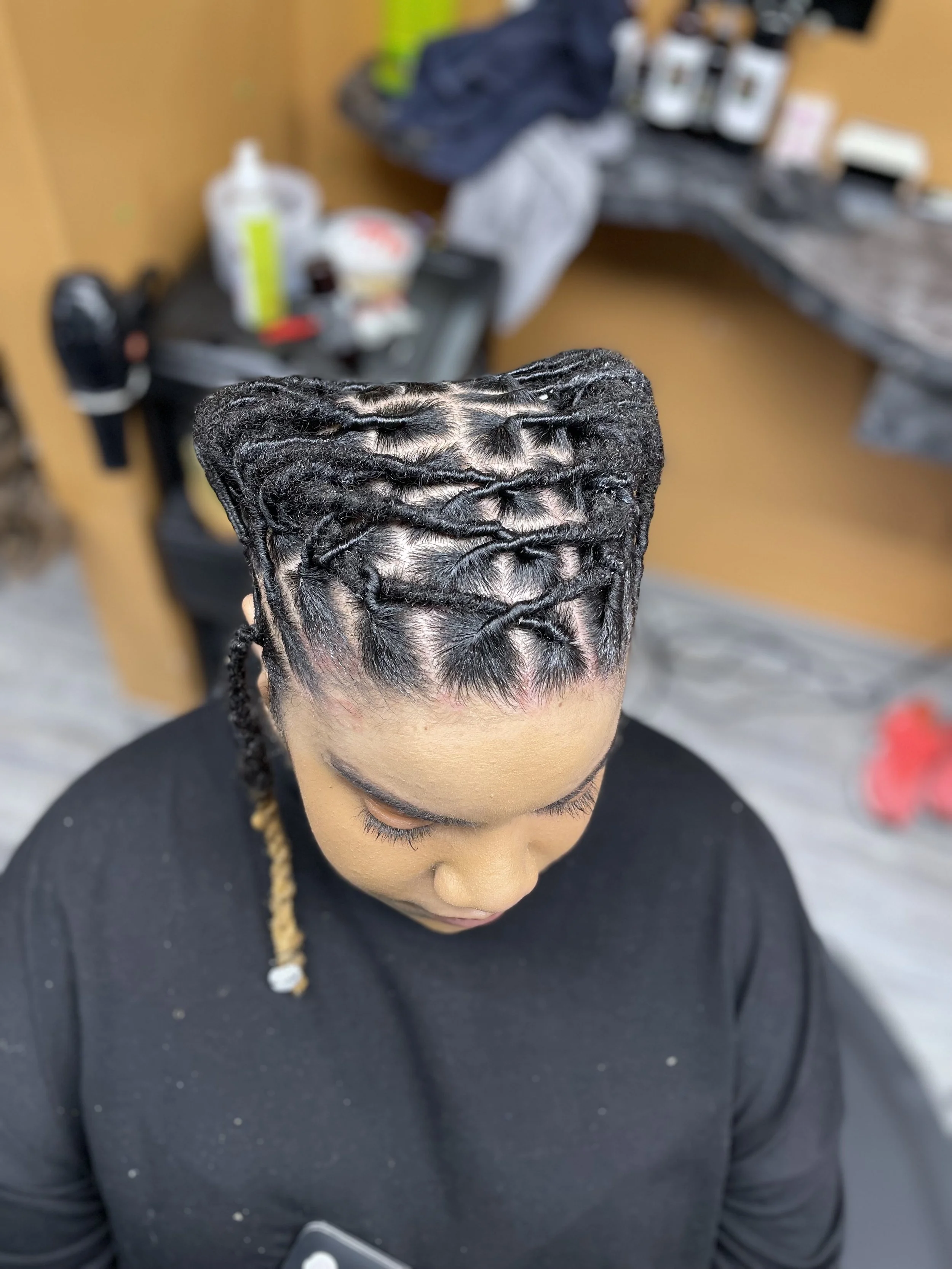 A woman with twisted braids in her hair, sitting in a salon chair with her head bowed down.