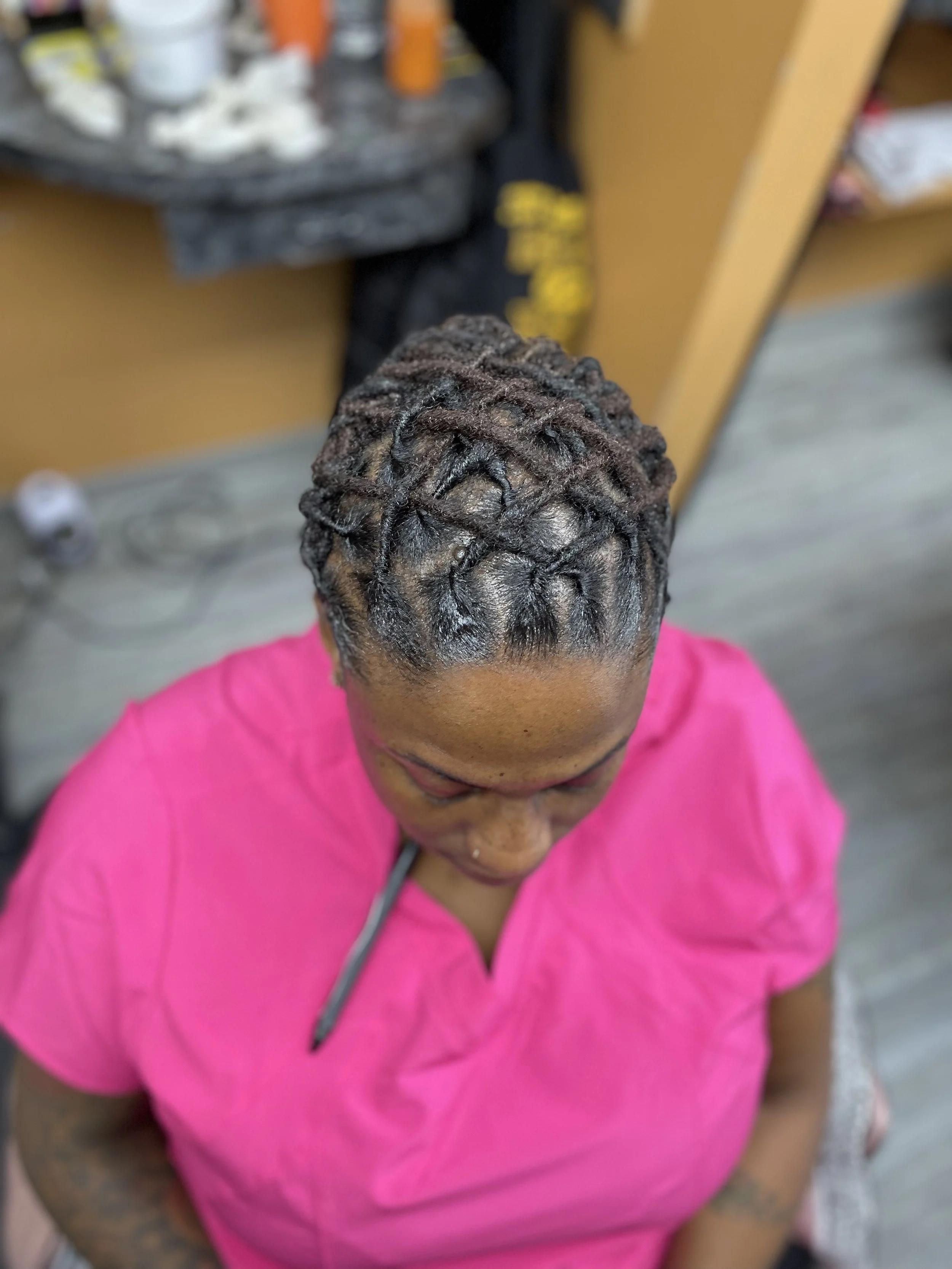 Woman with styled dreadlocks wearing a bright pink shirt, sitting in a salon.