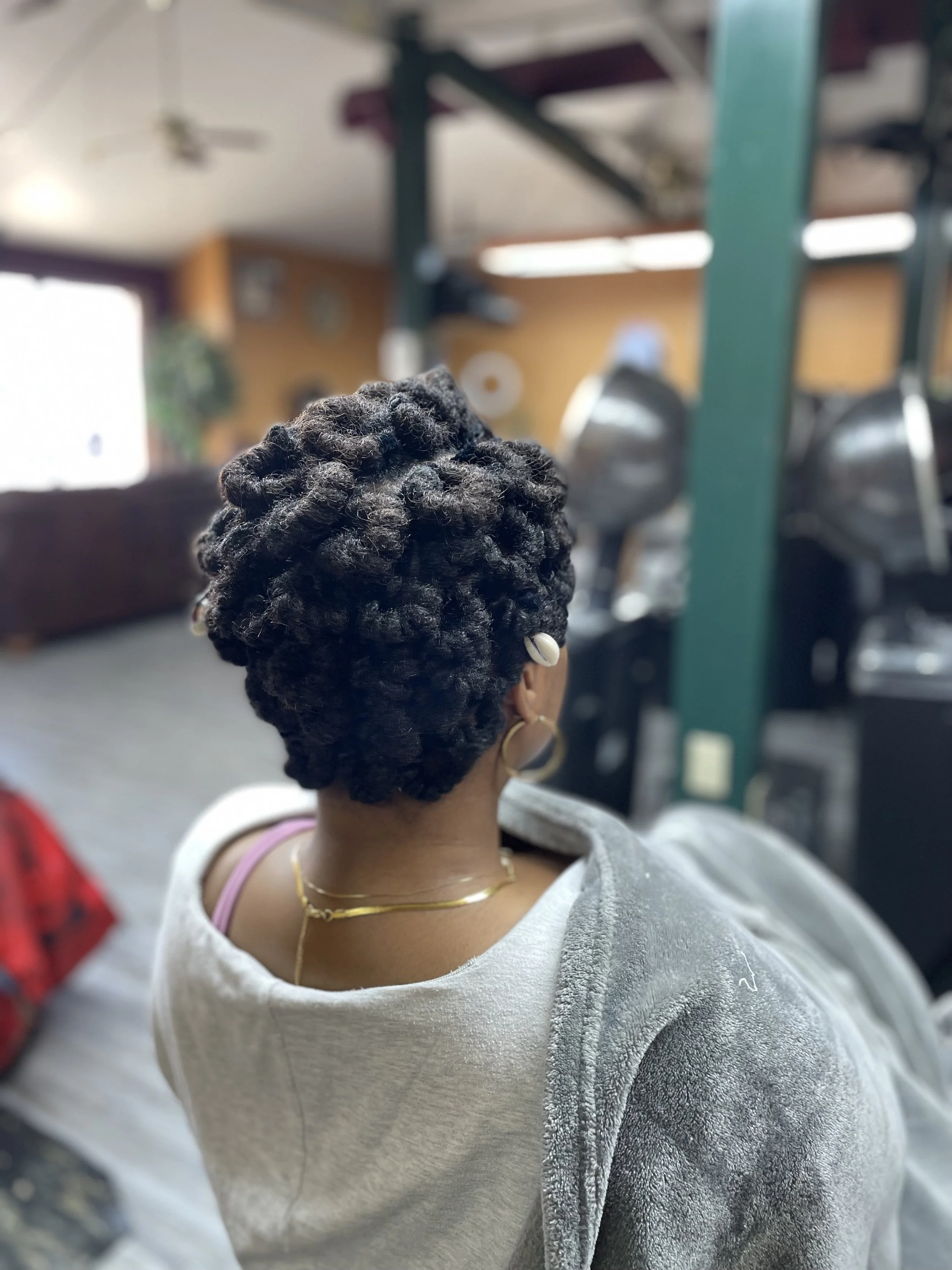 Back of a person with short, natural, curly hair styled in twists, wearing gold jewelry, seated indoors with a blurred background of kitchen or salon equipment.