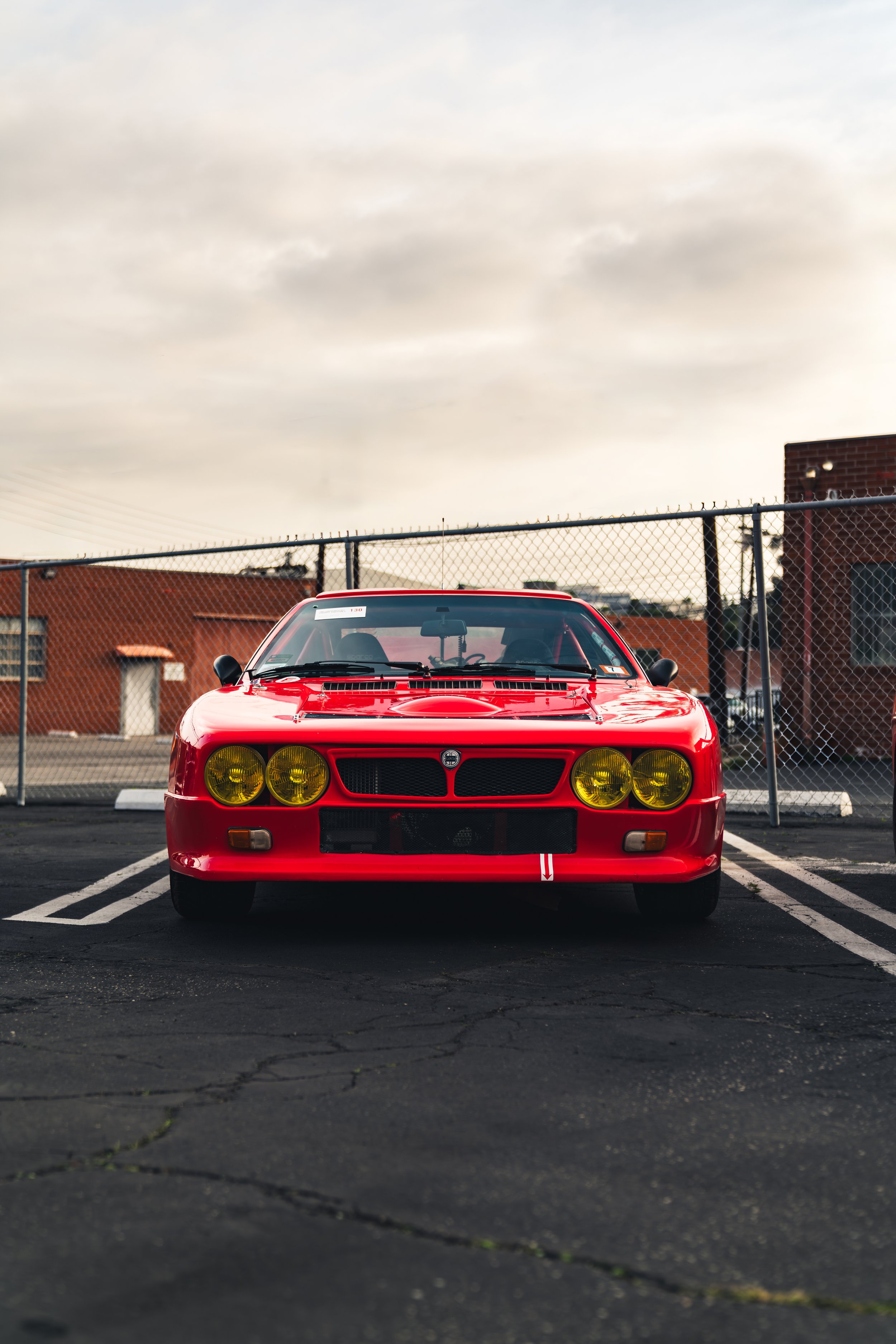 Front view of a red classic car with yellow headlights, parked in a lot with a wire fence and brick building in the background.