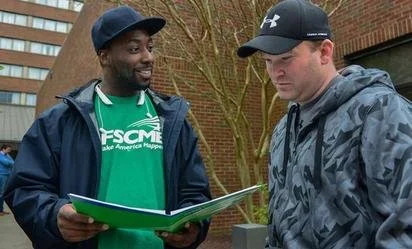 smiling AFSCME union member in green union T-shirt shows folder contents to white man with baseball cap