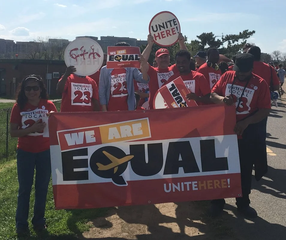 Group of Unite Here union members wearing red UH Local 23 tshirts holding up signs and large banner