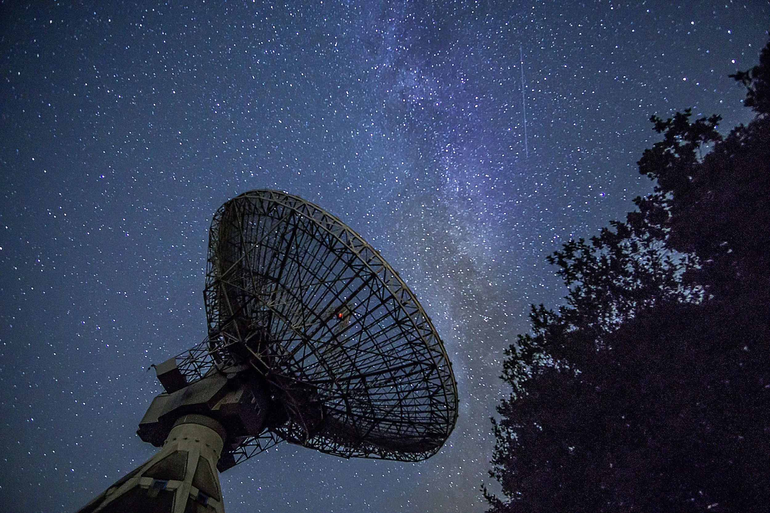 Night sky filled with stars and a visible Milky Way galaxy, with a radio telescope antenna in the foreground and trees on the right side.