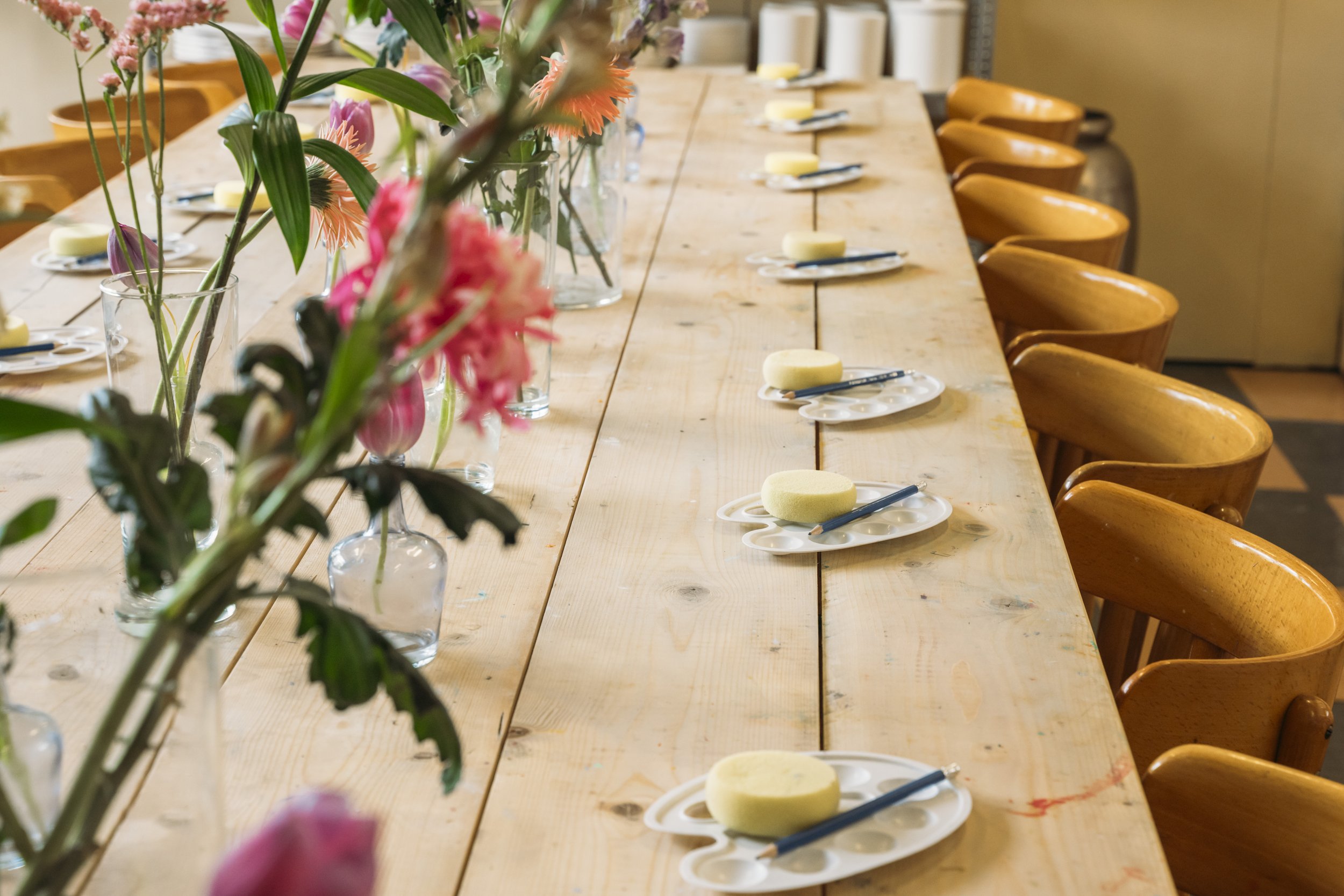 Een kunststudio met een vrouw die een boek leest, omgeven door schilderspullen, houten stoelen, en een bar met kopjes en borden, met bloemen en planten boven.
