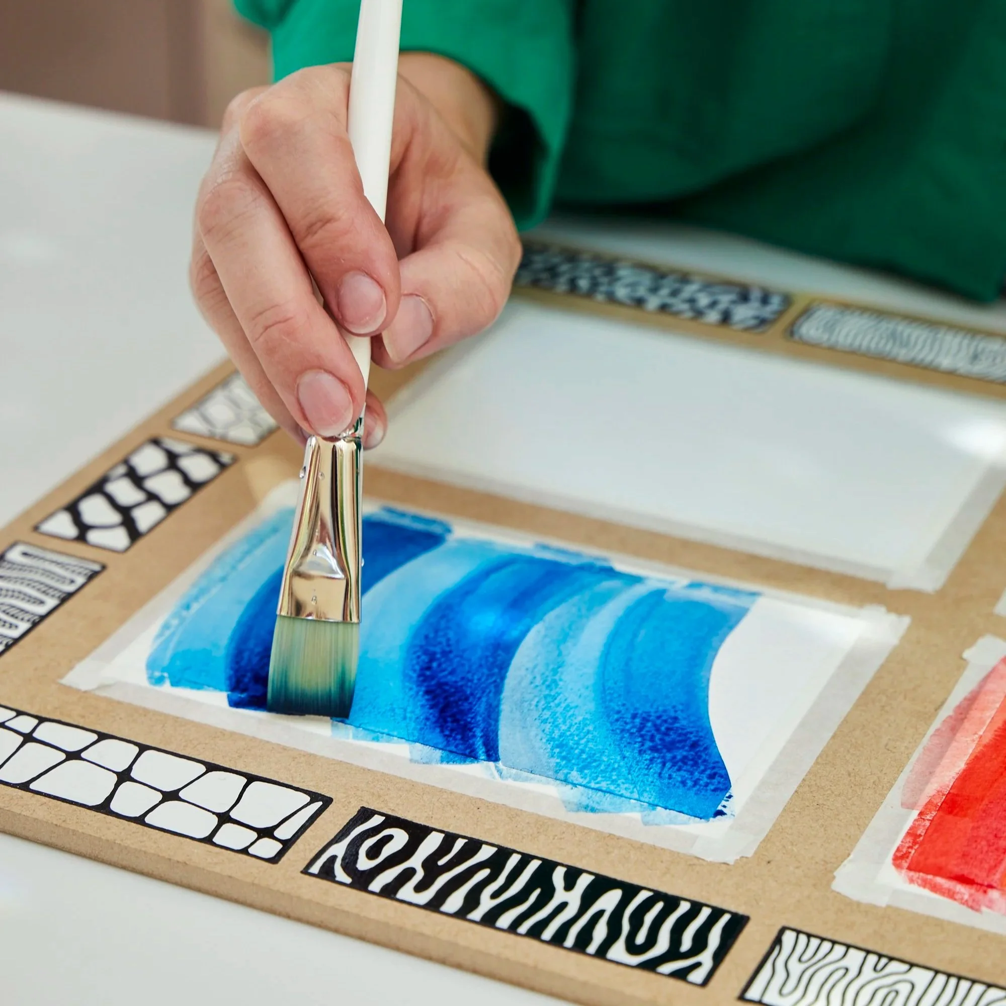 Close-up of a person's hand holding a paintbrush, painting a scene of blue ocean waves on a piece of paper on a cardboard surface.