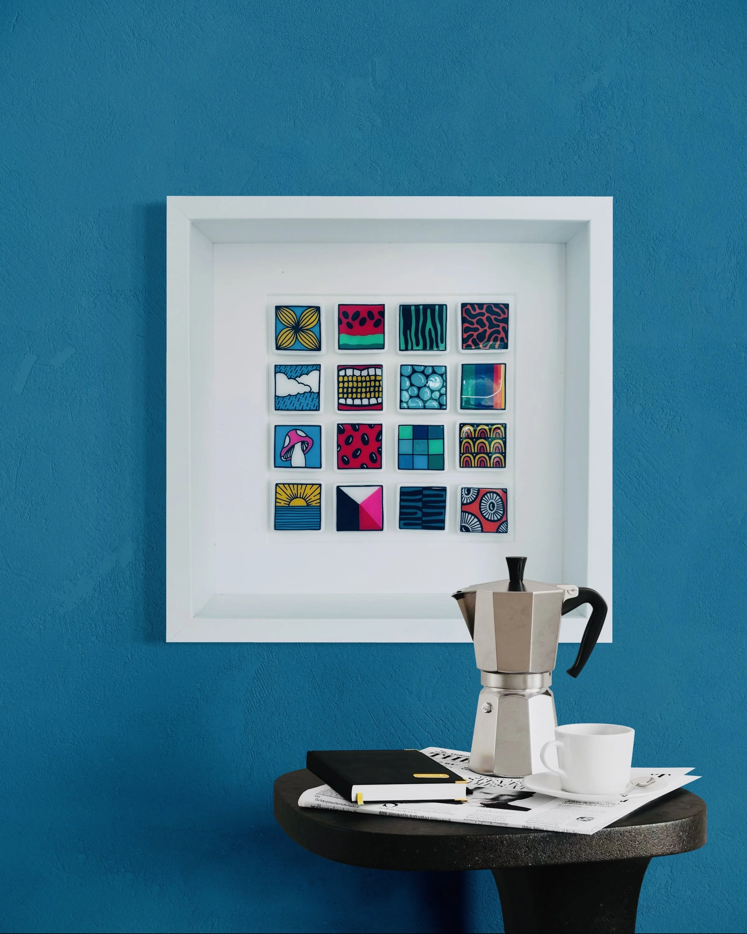 A white framed artwork with colourful abstract patterns hangs on a textured blue wall. The artwork contains small squares of various designs.