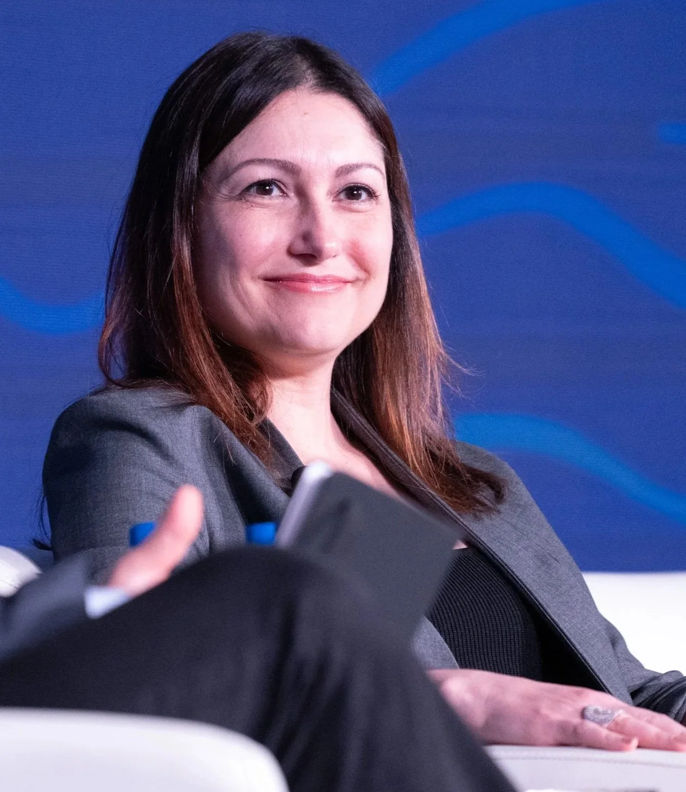 A woman with long brown hair and a gray blazer smiling during a speech or presentation with a blue background.